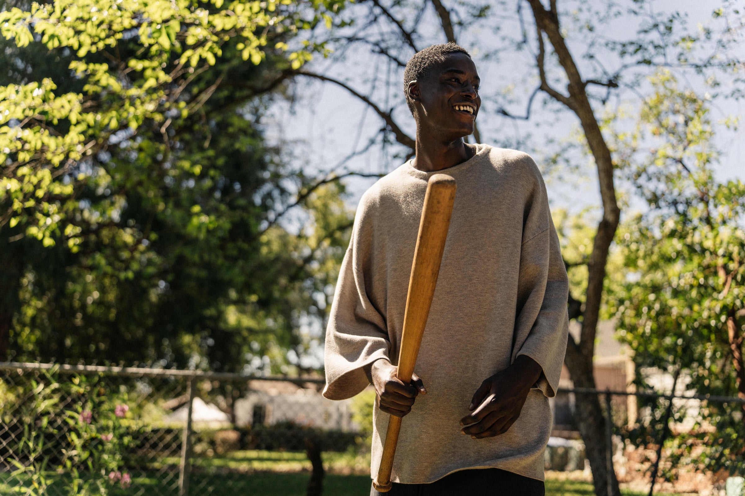 Young man standing outdoors in a sunlit yard wearing a loose beige sweater, holding a wooden baseball bat, with trees and a chain-link fence in the background.
