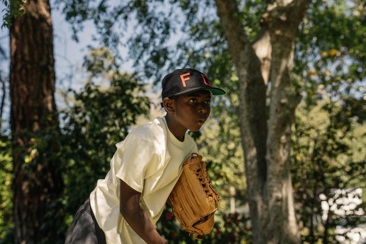 Boy wearing black cap and white shirt holding baseball glove outside among trees on a sunny day
