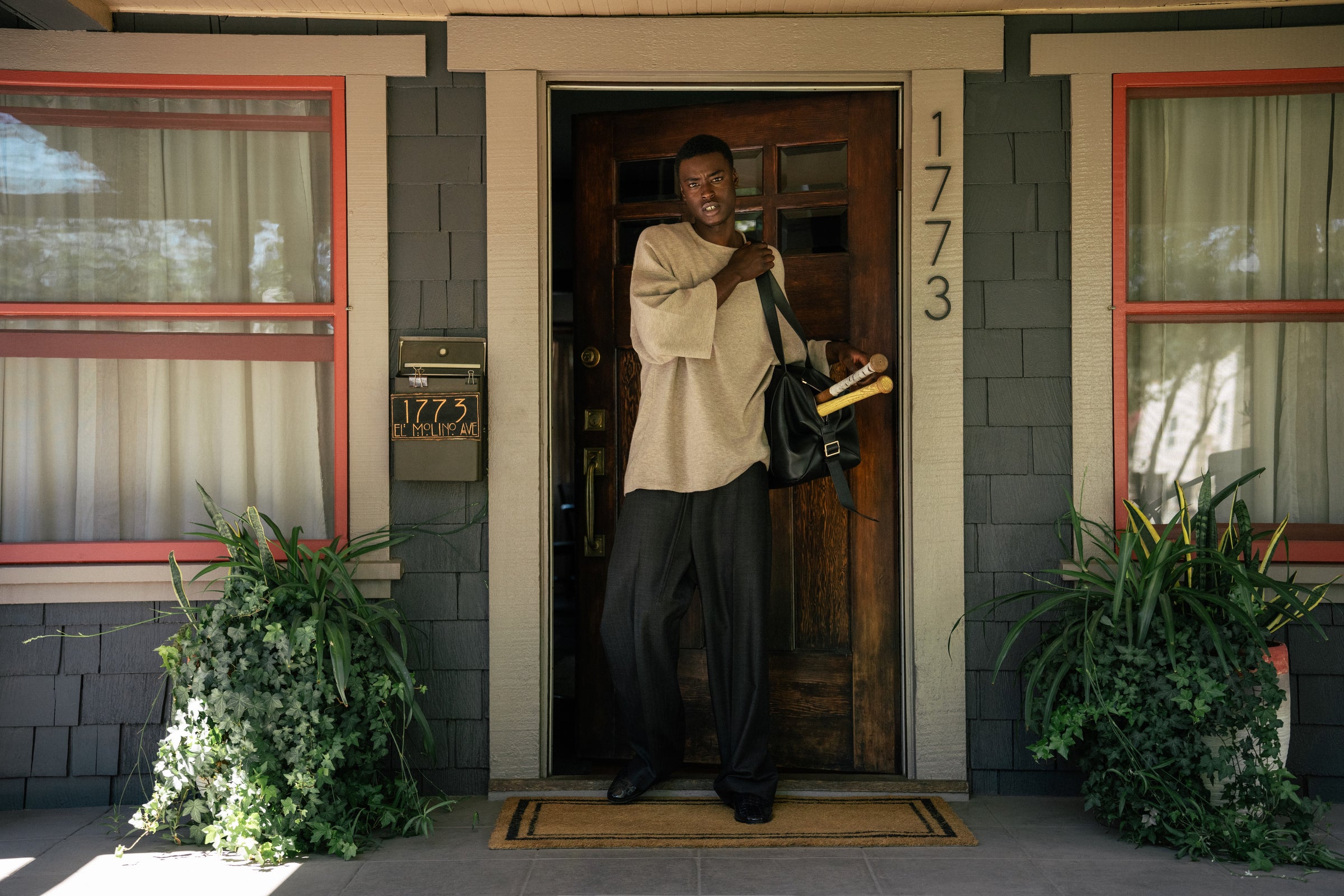 Man in beige sweater standing at wooden door of gray house holding bag and rolled papers by porch plants