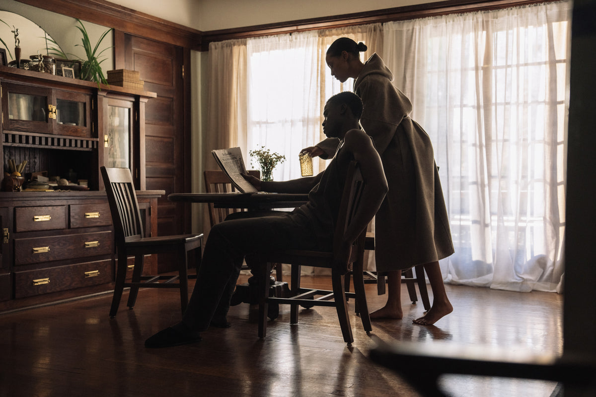 Two people interacting at a wooden dining table with sunlight streaming through sheer curtains in a cozy room