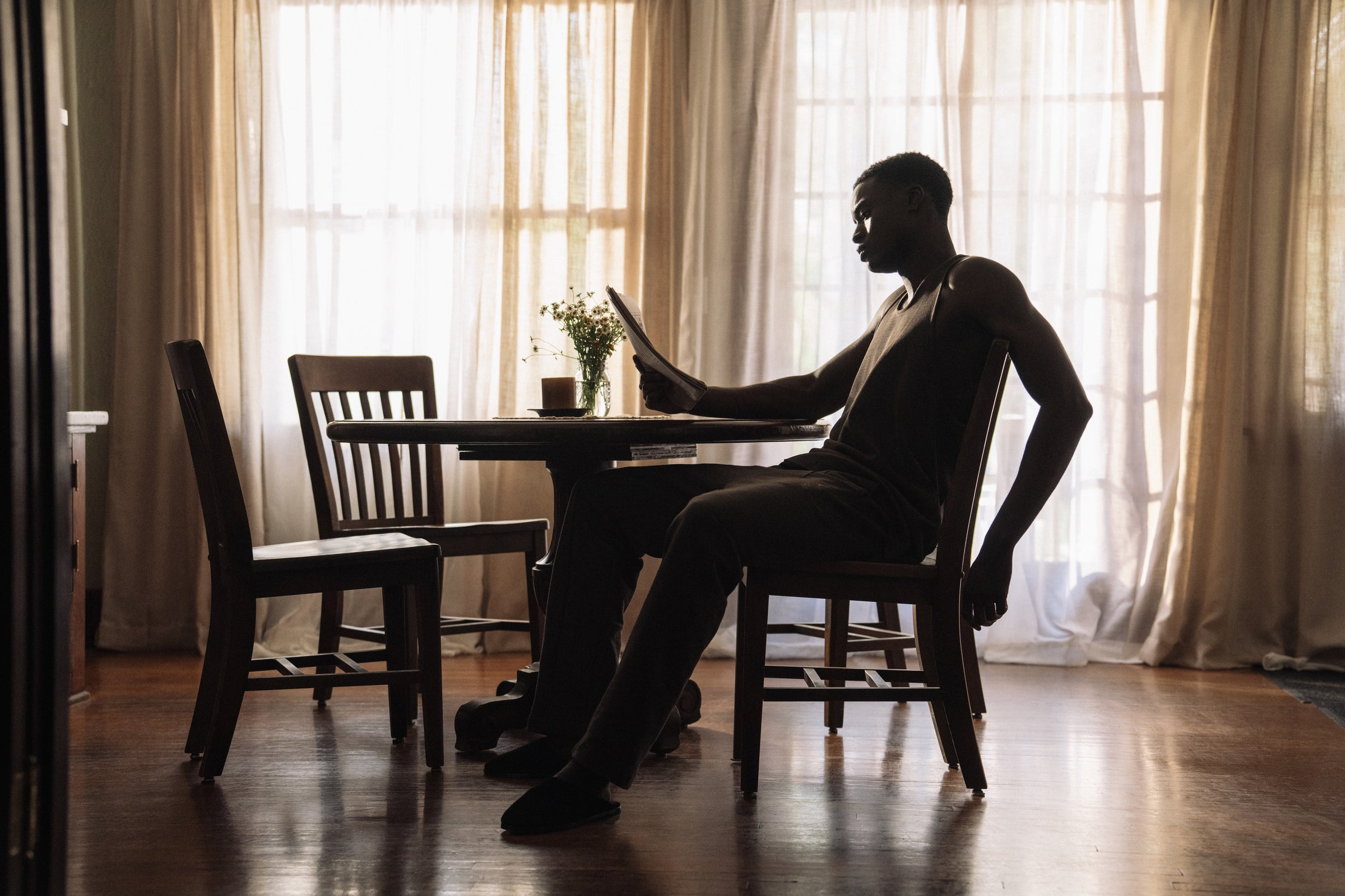 Silhouetted man reading a document while sitting at a round wooden table in a sunlit room with sheer curtains
