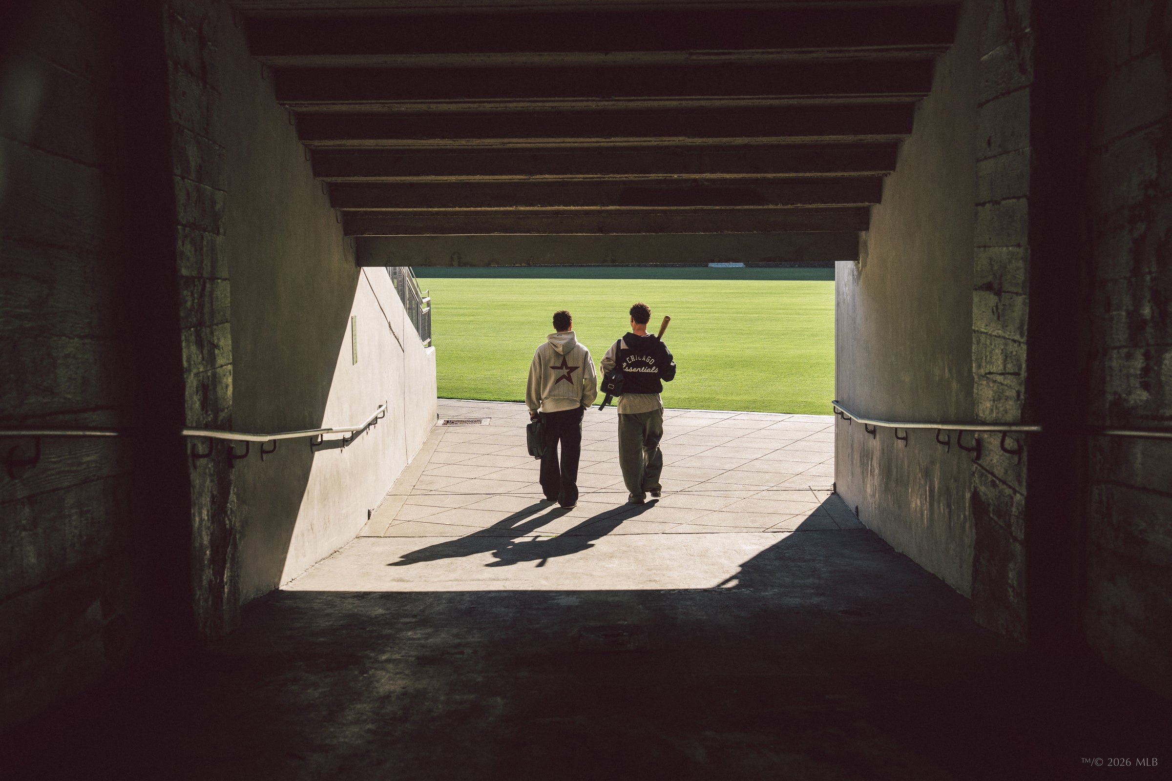 Two people walking through a shaded concrete tunnel toward a bright, grassy sports field in daylight