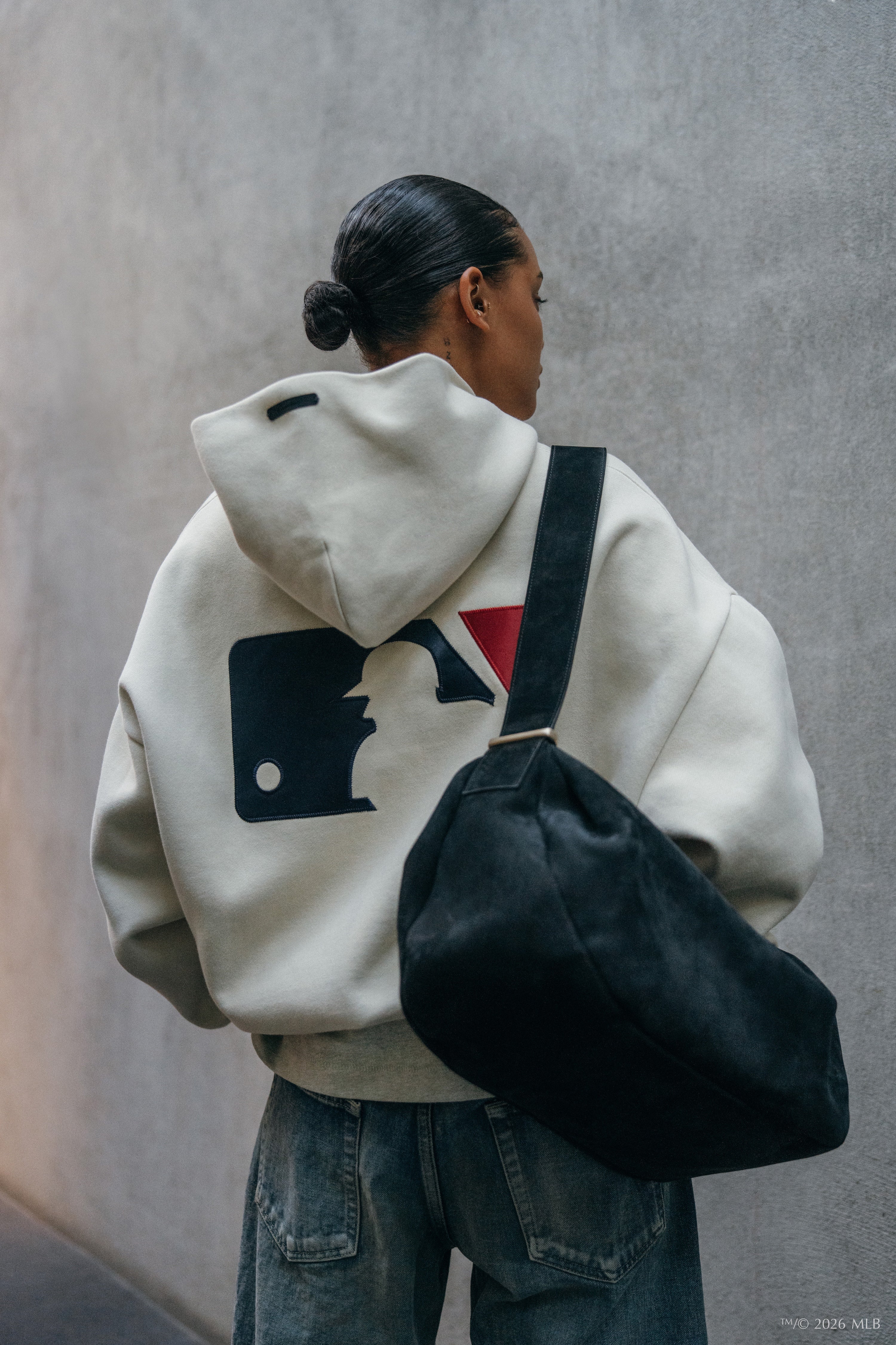Person wearing a cream hoodie with large navy and red graphic on back, carrying black shoulder bag against gray wall background