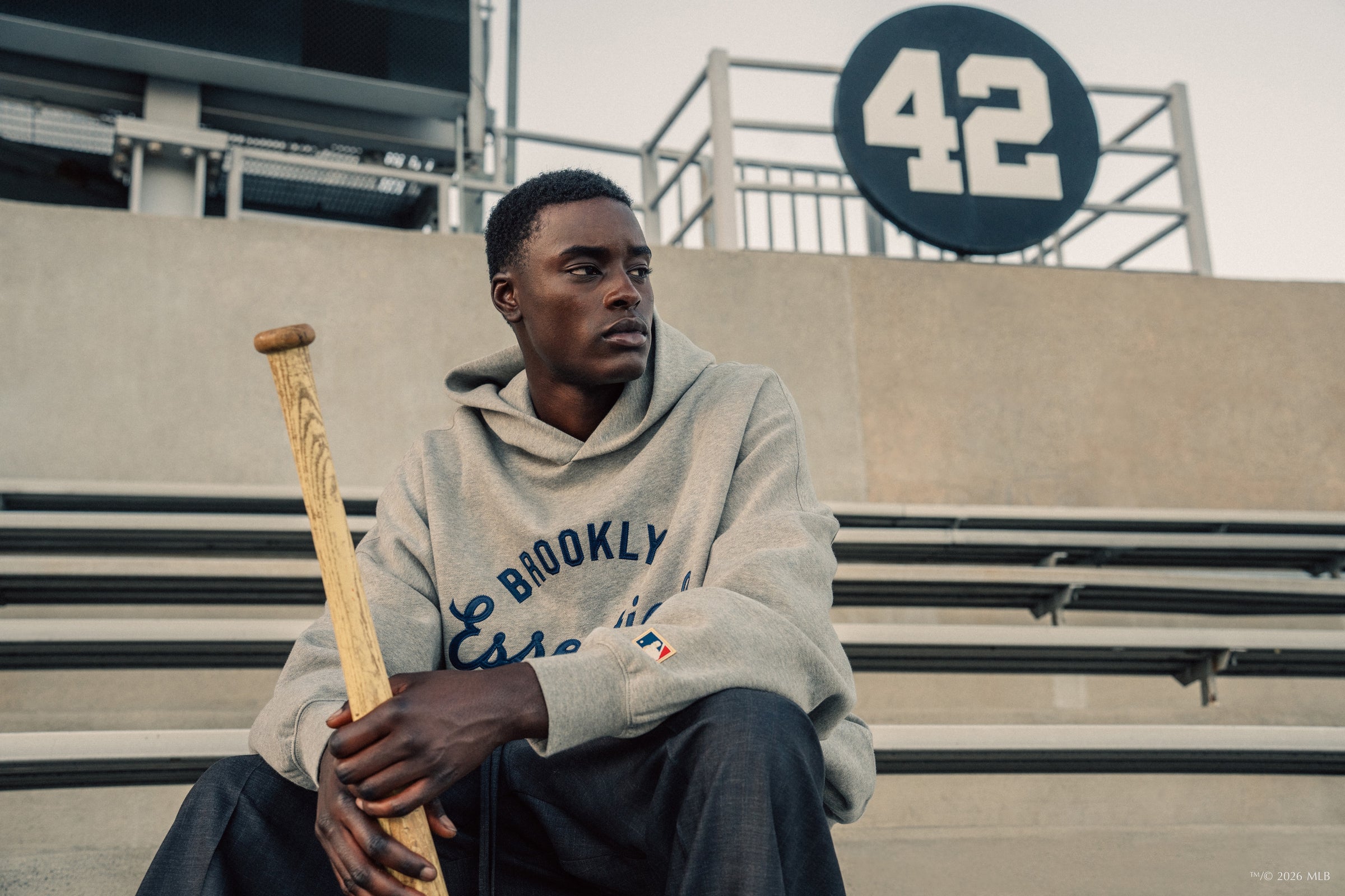 Man in gray hoodie holding wooden baseball bat sitting on stadium bleachers with number 42 sign behind him