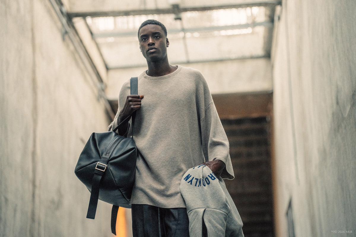 Young man in loose beige sweater holding bag and gray Brooklyn sweatshirt walking through industrial stairwell