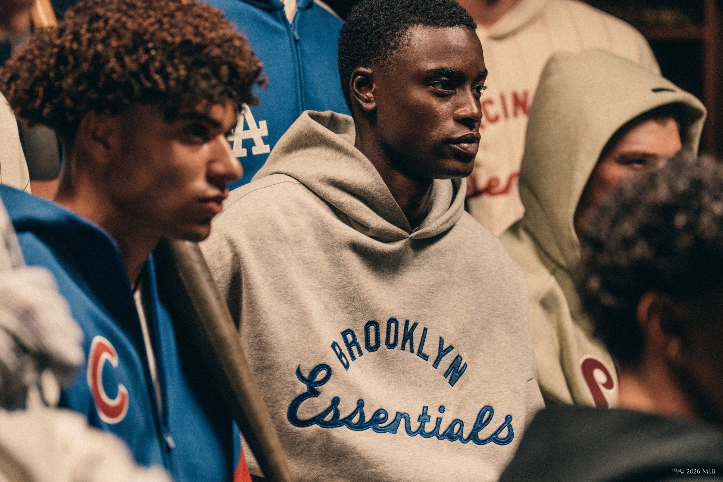 Group of young men in varied hoodies, one wearing gray Brooklyn Essentials sweatshirt, engaged indoors with focused expressions