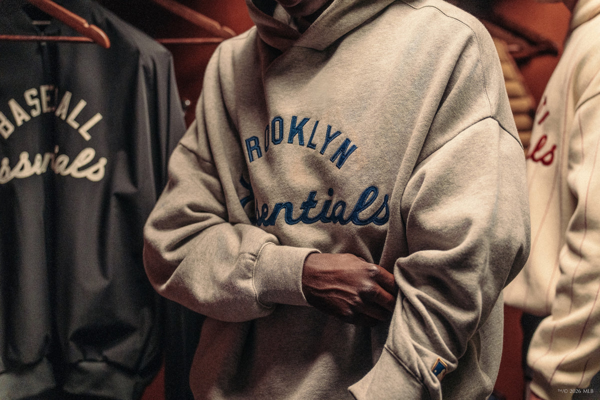 Person wearing light gray Brooklyn Essentials hoodie standing among hanging vintage baseball jackets
