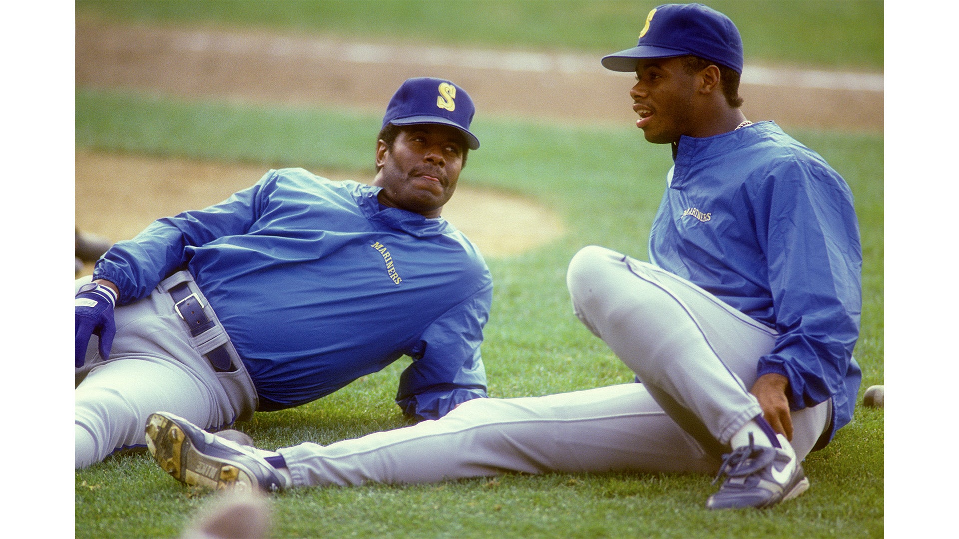 Two baseball players in blue warm-up jackets and caps sit on the grass stretching before a game on a daytime field.