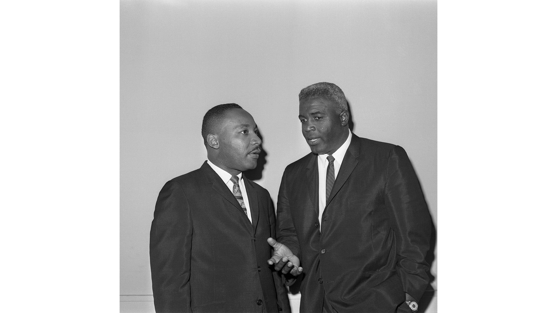 Black and white photograph of two men in dark suits and ties engaged in conversation against a plain background, one gesturing with his hand while the other listens attentively.