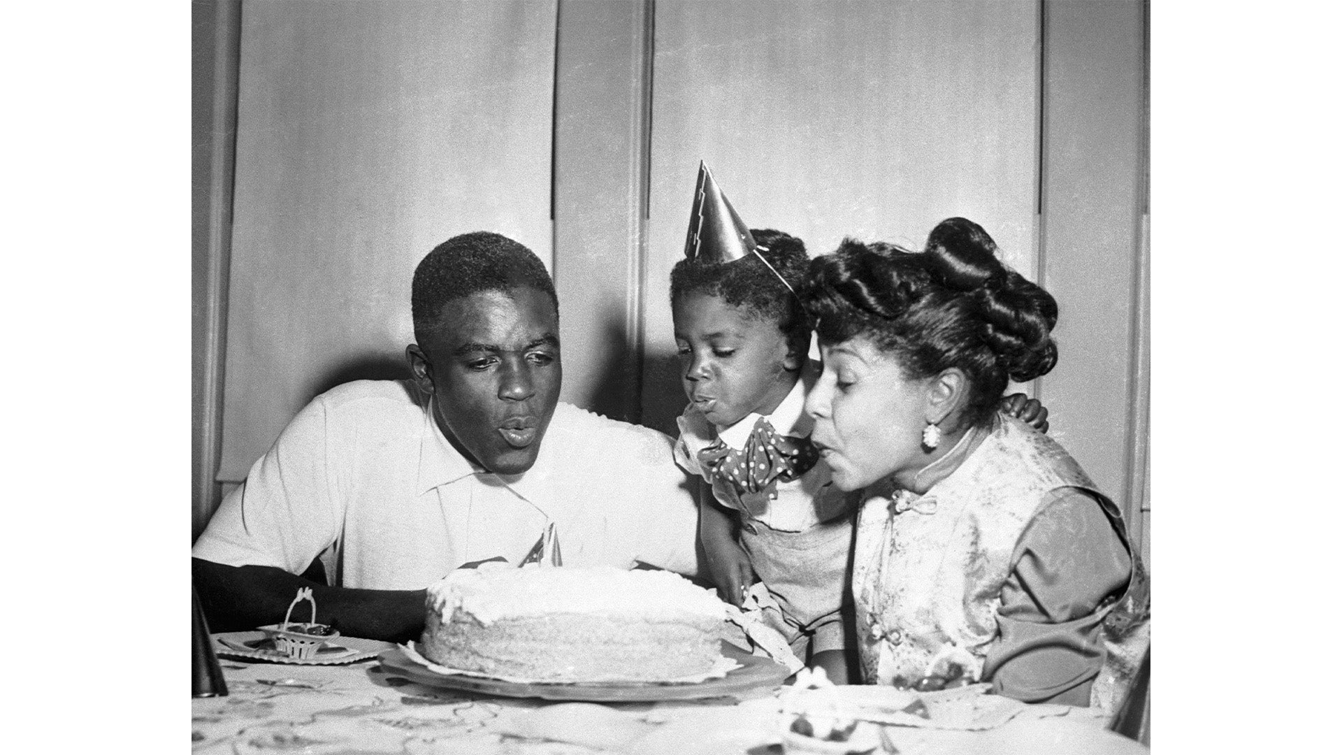 A black and white photograph of three people gathered around a table, blowing out candles on a birthday cake, with two women and one man smiling and leaning forward.