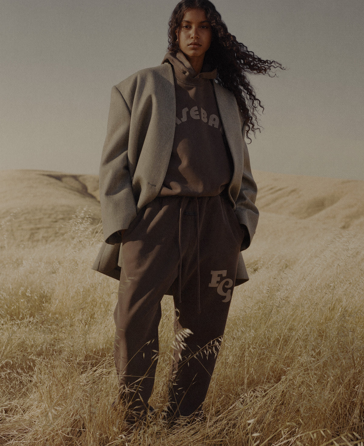 Woman wearing a gray blazer over a dark hoodie and matching trousers standing in tall golden grass with rolling dunes behind her under a muted sky