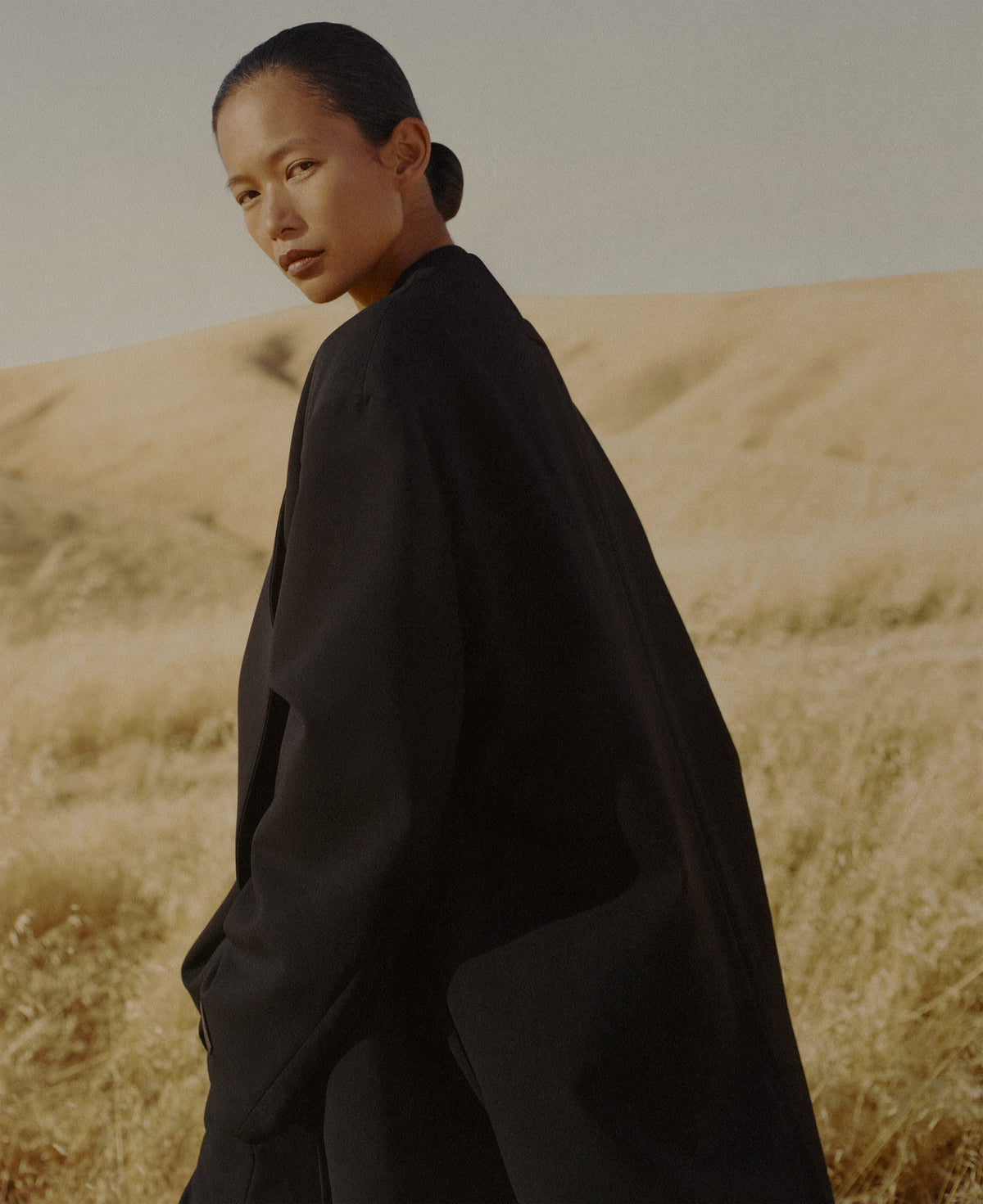 Woman with tied back hair wearing flowing black Outerwear and looking over her shoulder in a sunlit golden grass field beneath a muted sky