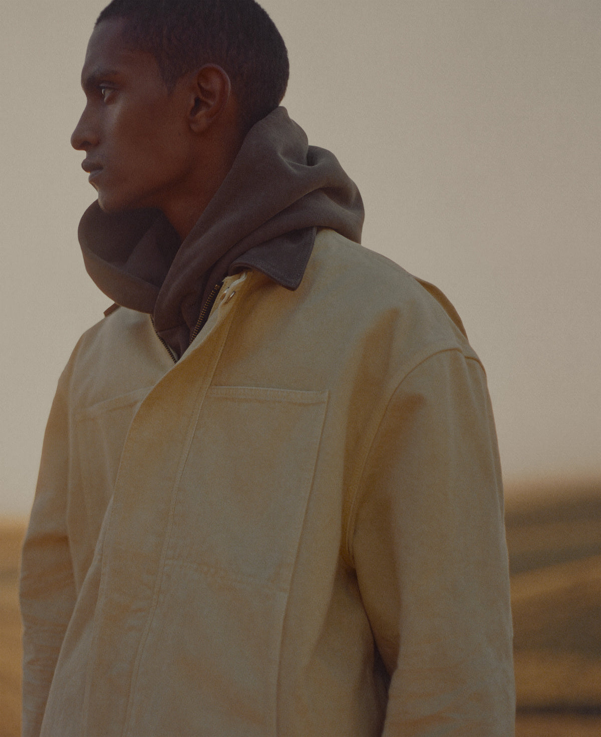Profile portrait of a man in a pale yellow Outerwear jacket layered over a dark hooded top against a softly blurred sunlit landscape