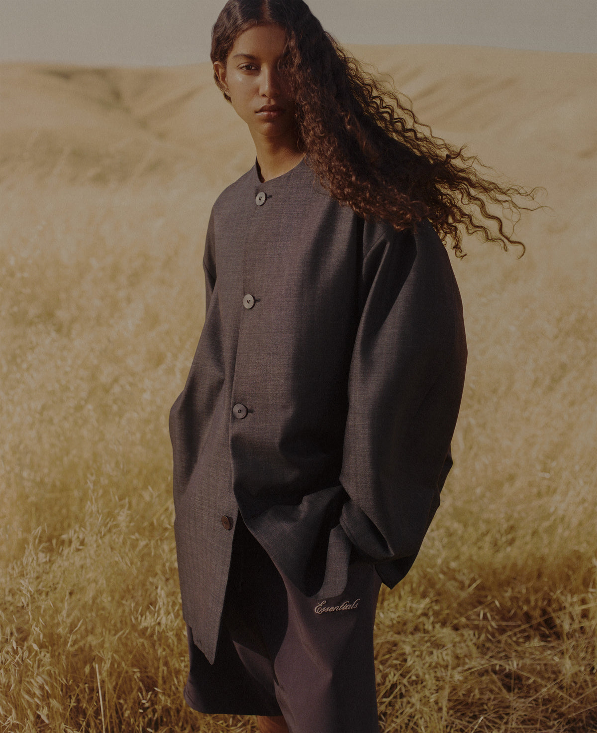 Woman with windblown curly hair wearing oversized dark buttoned Outerwear while standing in a sunlit dry grass field beneath softly rolling hills