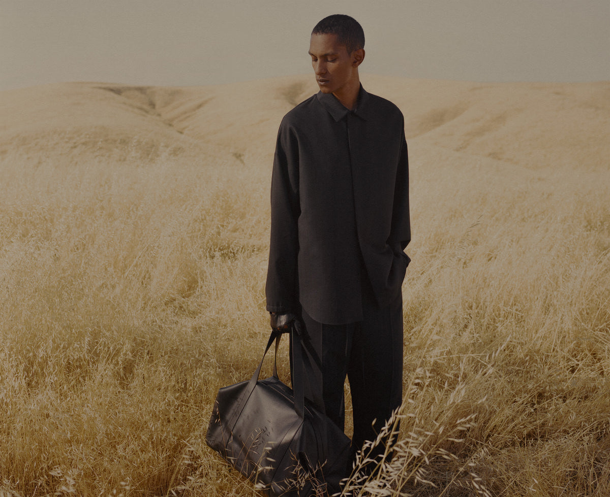 Man in a dark shirt and trousers carrying a leather duffel bag through tall golden grass with rolling hills beneath a muted sky