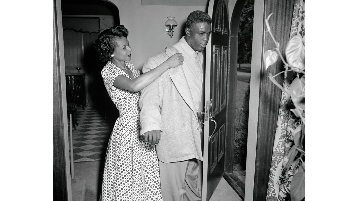 A black and white photograph of a woman in a patterned dress helping a man put on a coat inside a home near the front door, with a glimpse of a checkered floor in the background.