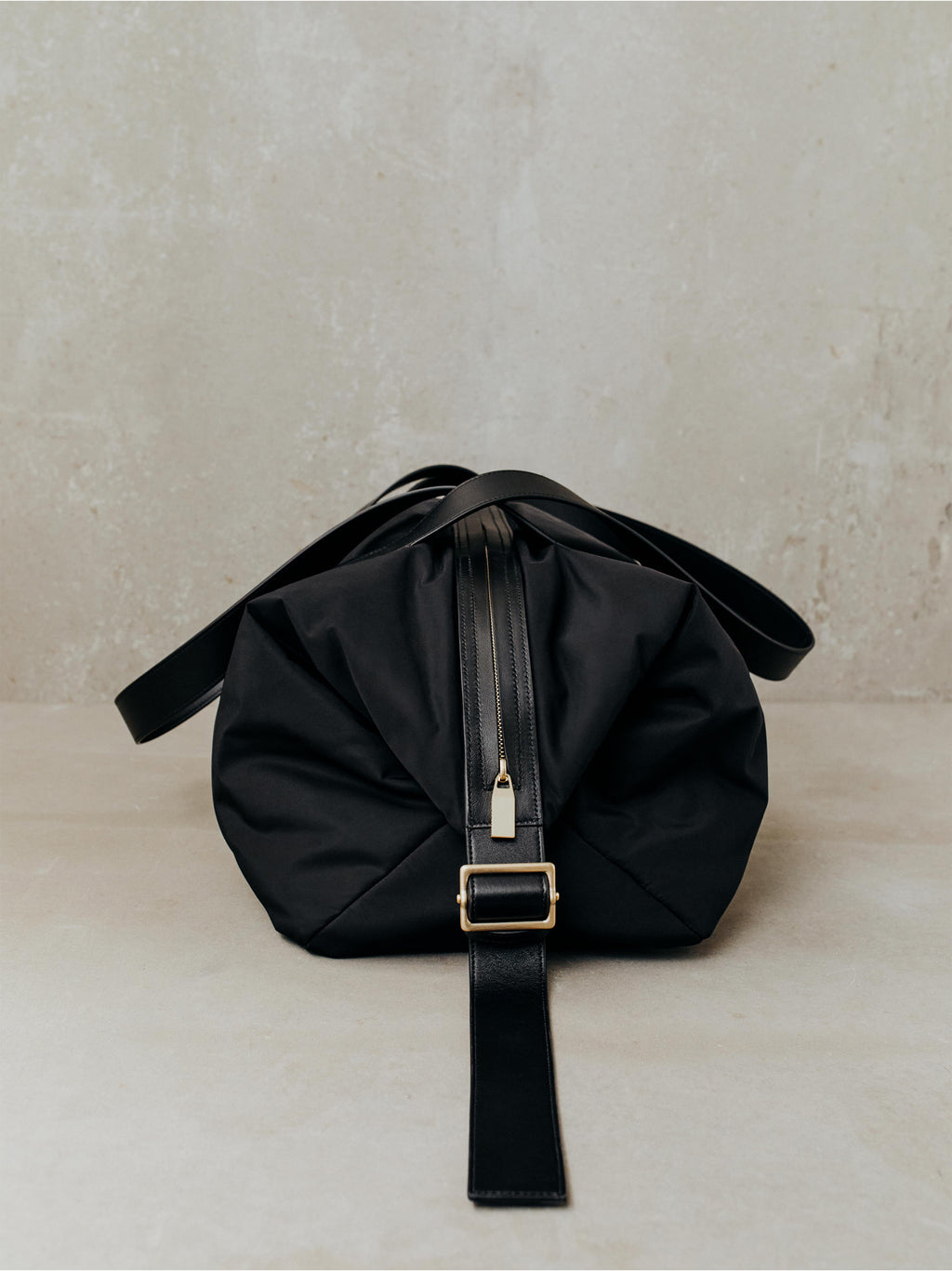 Black nylon duffel bag with central zipper, leather strap, and gold-tone buckle on neutral background