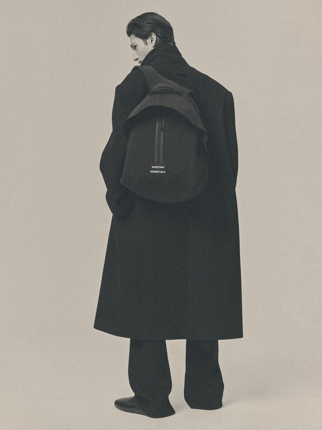Essentials Backpack in black worn by a model over a long dark coat showing its rounded silhouette and small logo against a muted neutral studio background
