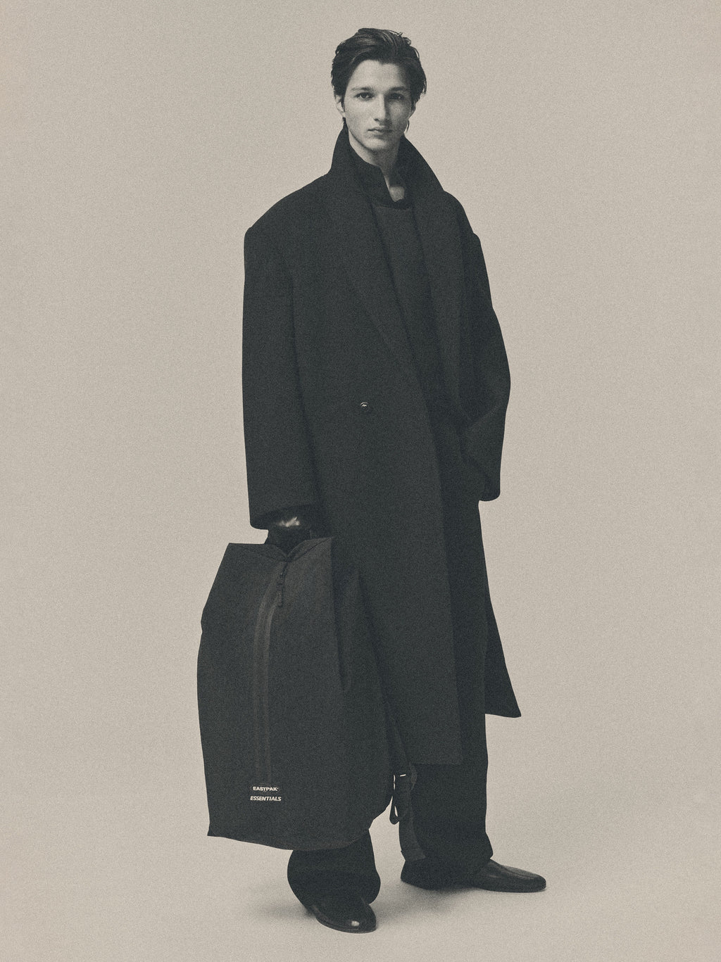 Sports Duffel in black carried by a model in a long dark coat showing its tall structured shape padded handles and spacious design against a neutral studio background
