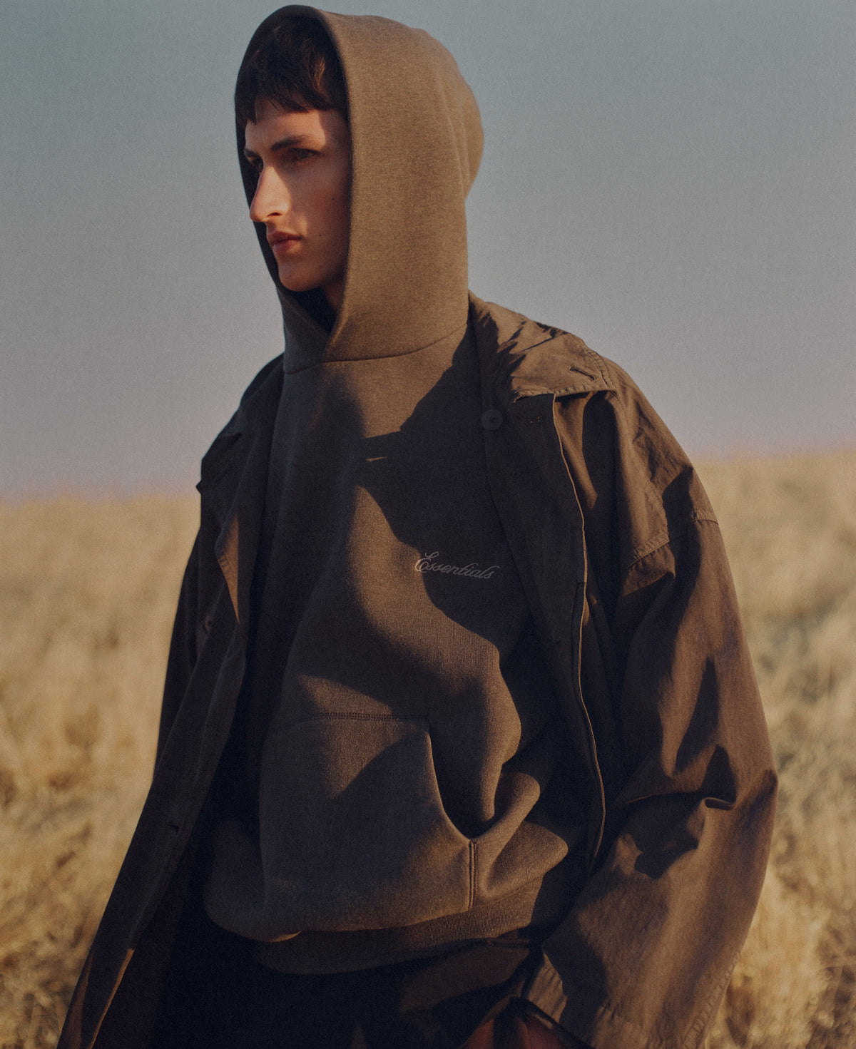 Young man wearing a dark hooded top and lightweight outerwear standing in a sunlit field of tall dry grass under a muted sky