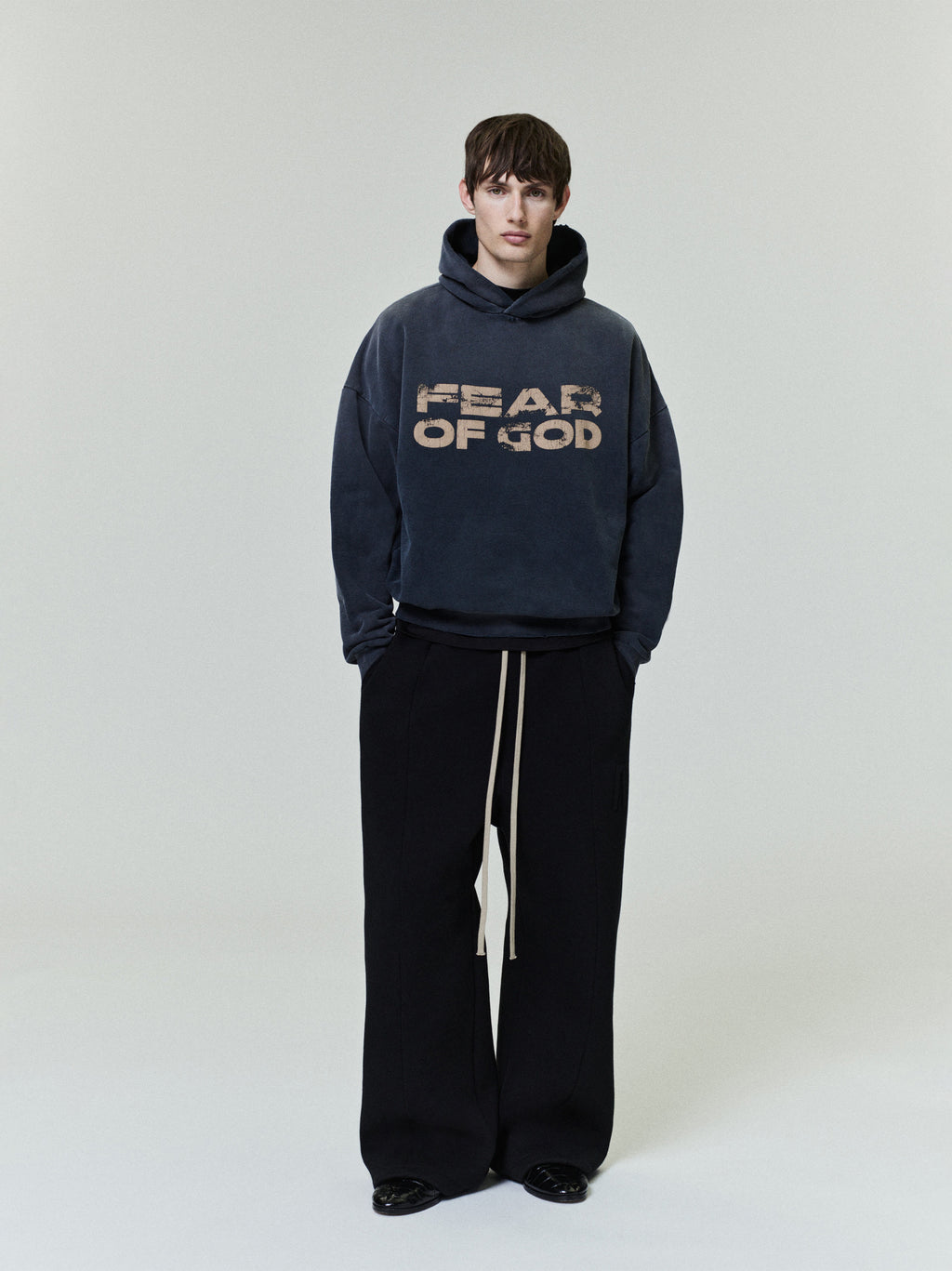 model wearing faded navy cotton fleece hoodie with distressed "FEAR OF GOD" graphic and wide-leg black pants