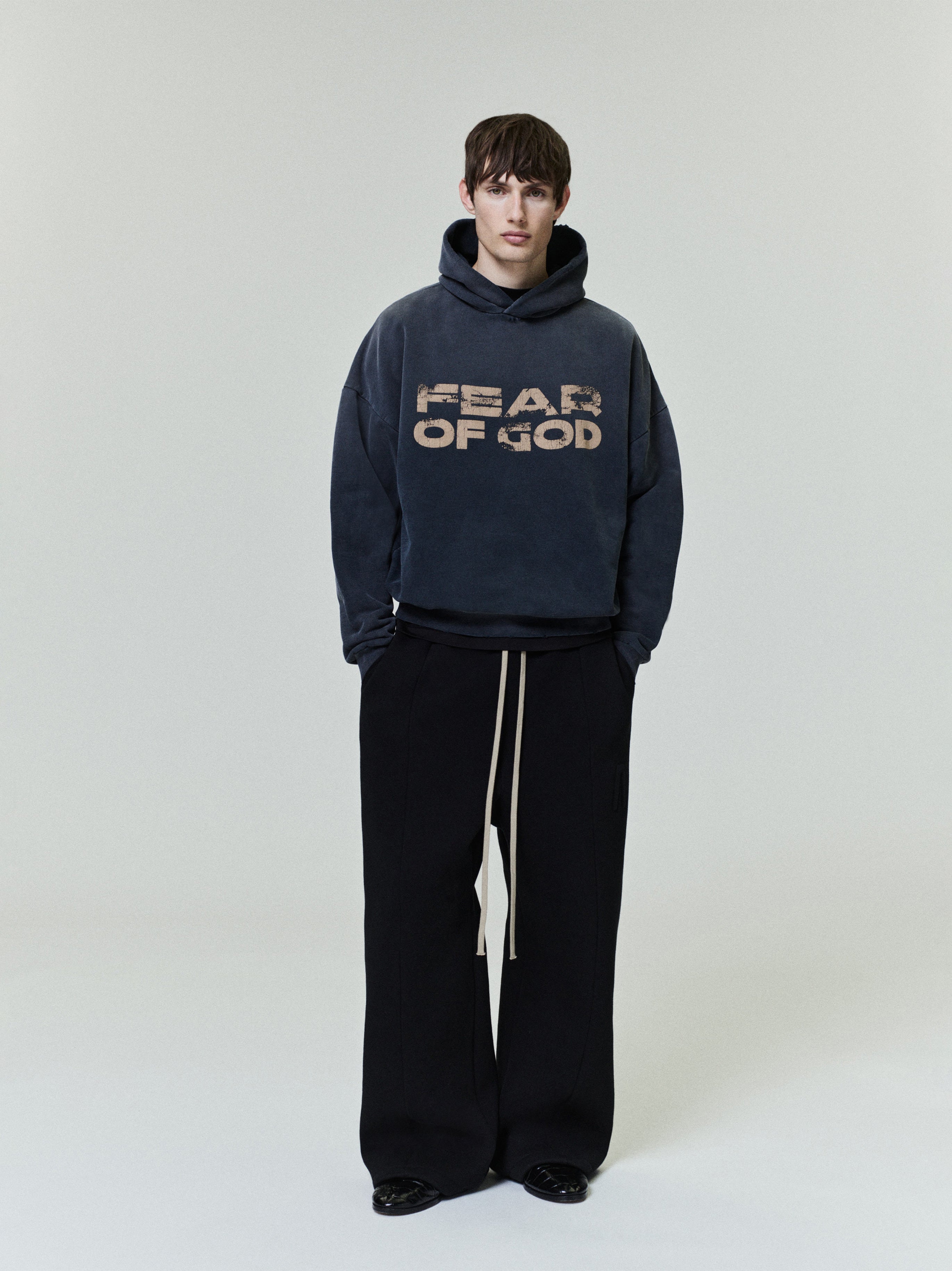 model wearing faded navy cotton fleece hoodie with distressed "FEAR OF GOD" graphic and wide-leg black pants