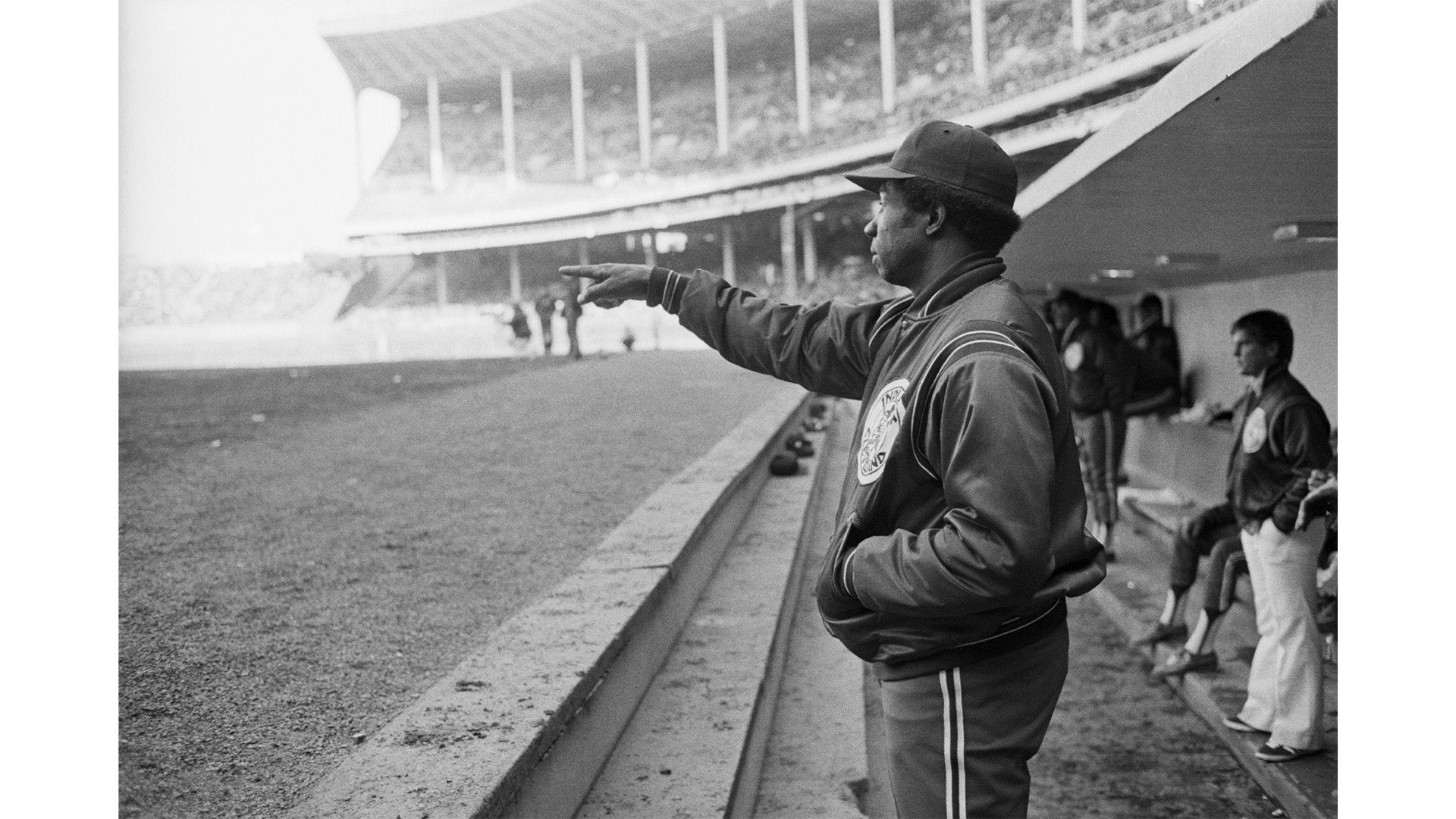 A black and white photograph of a baseball coach or player in a jacket and hat standing near a dugout railing pointing toward the field, with teammates sitting and standing behind him.