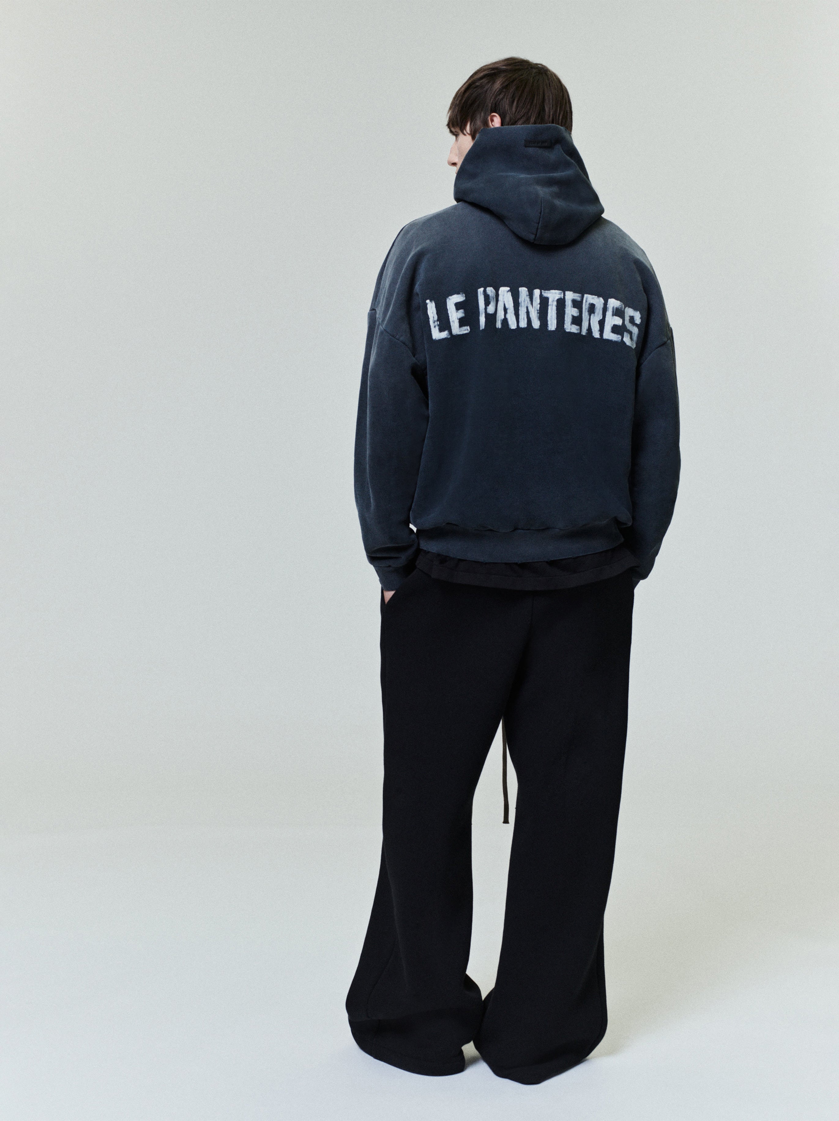 Le Panteres Hoodie in washed black with bold distressed lettering across the back, paired with loose-fitting black trousers for a modern casual outerwear look.