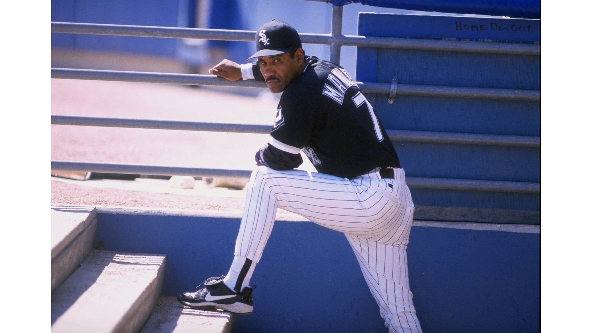 A baseball player wearing a black jersey with the number 7 and white pinstriped pants stretches one leg on a step near a dugout railing while looking toward the camera.