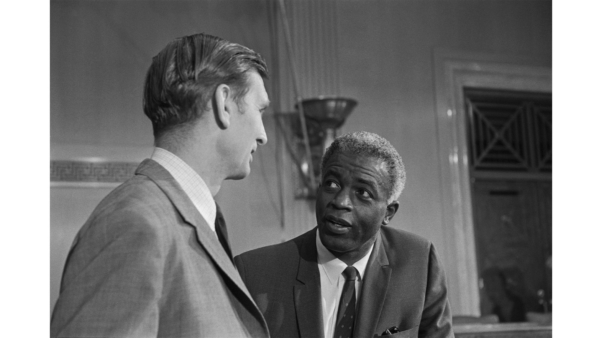 A black and white photograph of two men in suits engaged in conversation in an indoor setting, with one man looking attentively at the other.