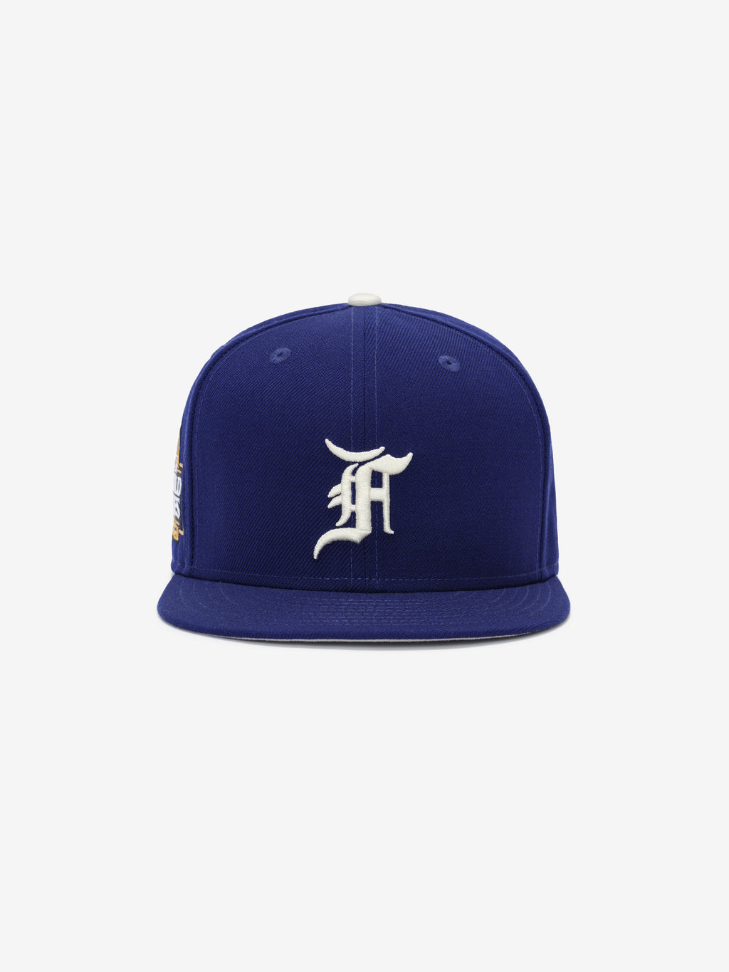 Front view of a royal blue fitted cap with a raised white ornate letter embroidered on the front panel