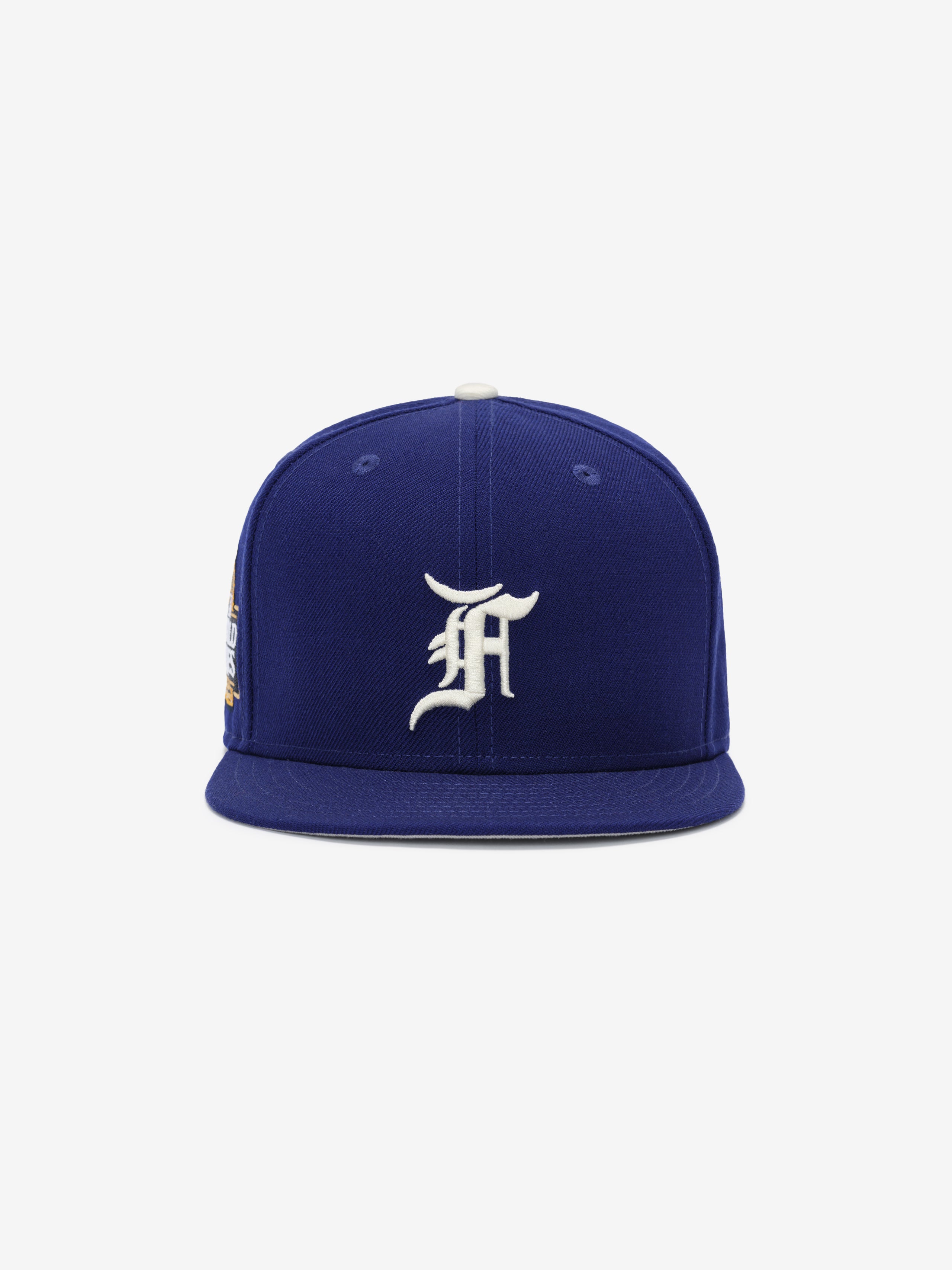 Front view of a royal blue fitted cap with a raised white ornate letter embroidered on the front panel