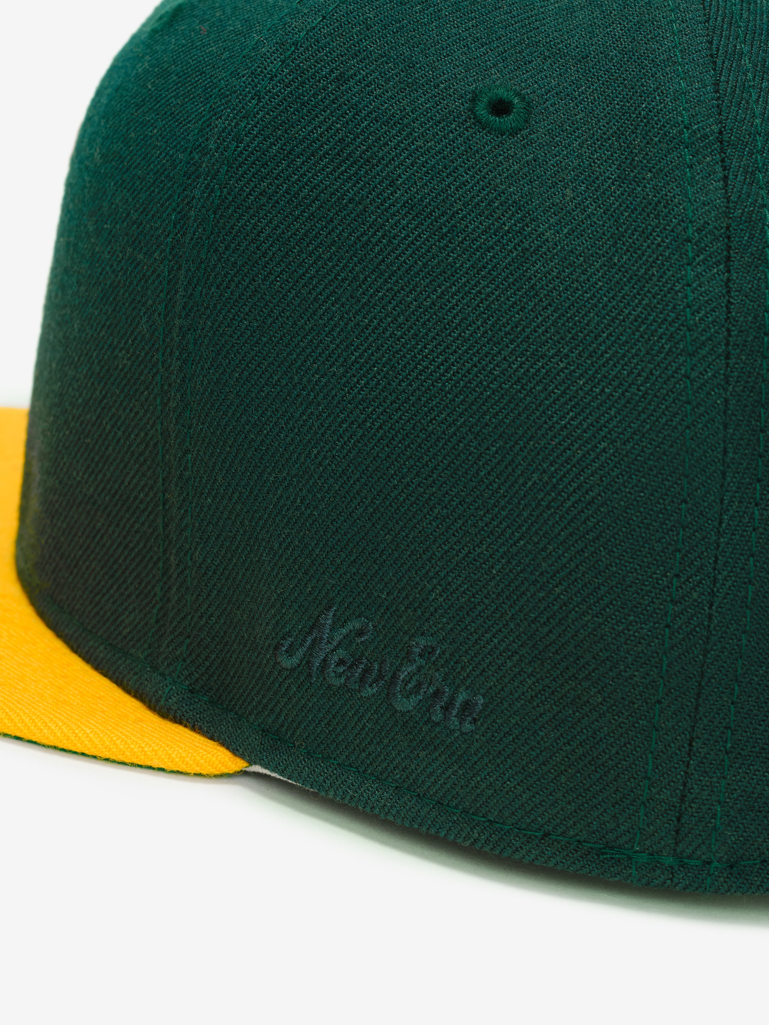 Fear of God Essentials 59FIFTY Athletics Cap close-up showing a dark green fabric crown with subtle tonal embroidery and a bright yellow flat brim.
