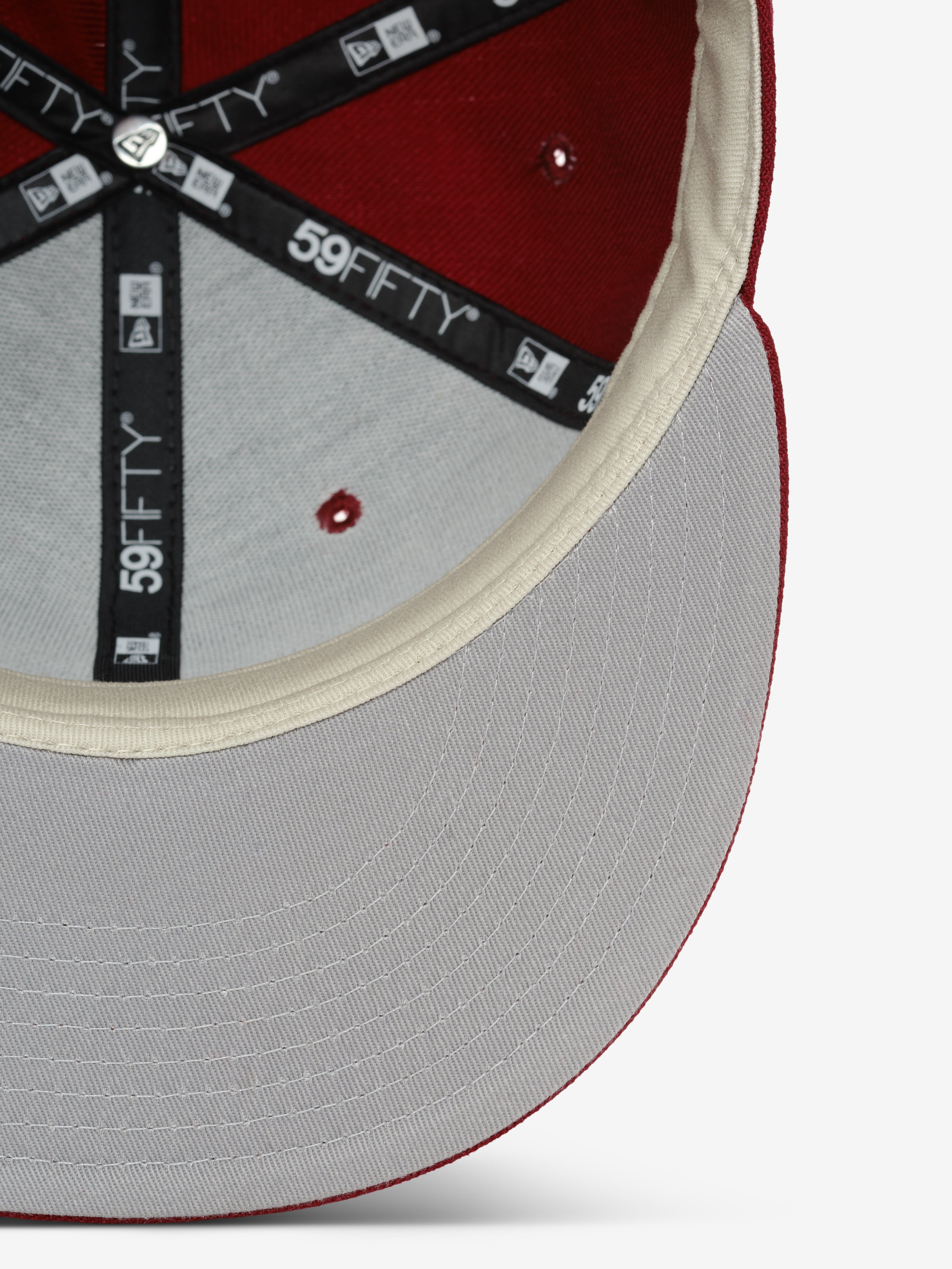 Fear of God Essentials 59FIFTY Athletics Cap shown from the inside, featuring a cream-colored inner sweatband, black branded taping with 59FIFTY logos, and a light gray underside brim contrasted by a rich crimson crown.