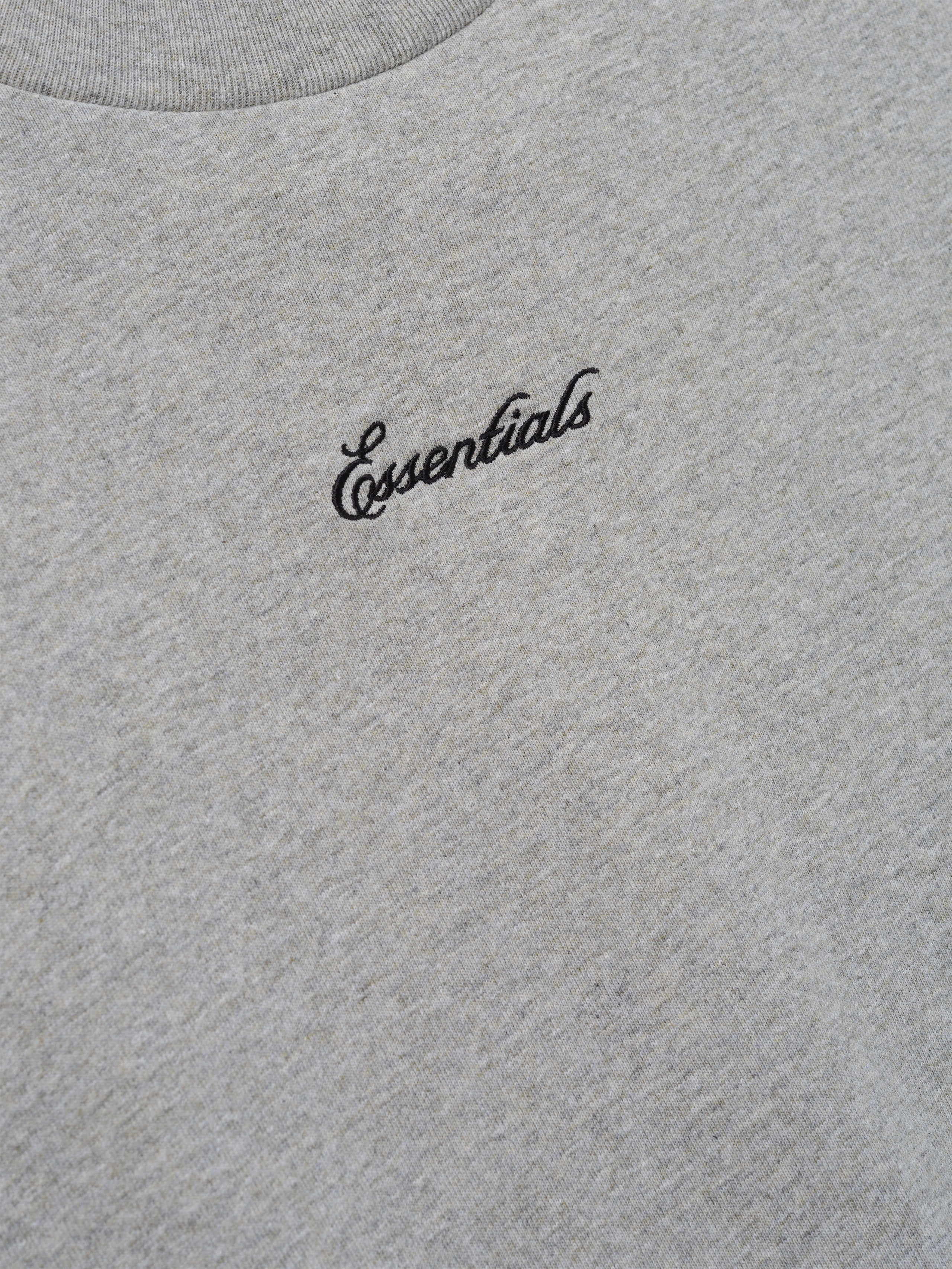 Close-up of the Kids Signature 90s Tee in soft concrete heather fabric featuring black cursive Essentials embroidery on the chest for a casual, classic look.
