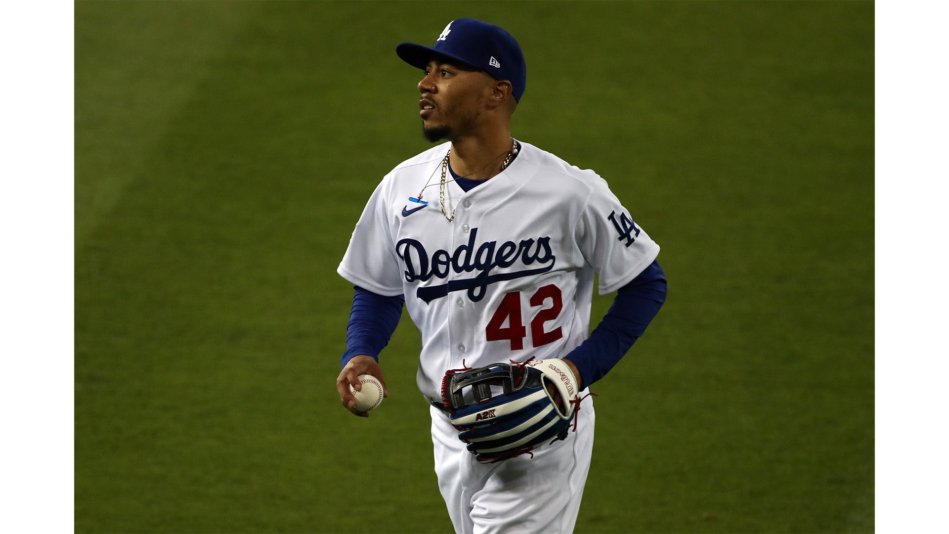 A baseball player wearing a white Los Angeles Dodgers uniform with the number 42 stands on the field holding a baseball and glove, looking to his left during a game.