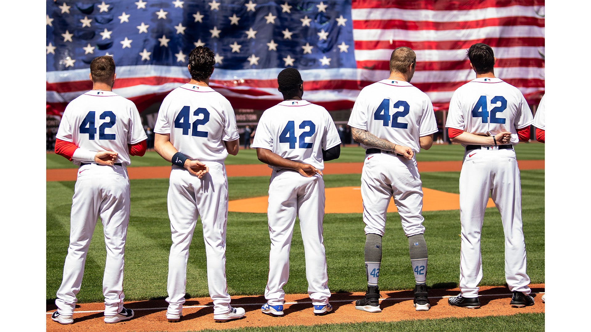 Five baseball players stand side by side facing a large American flag, each wearing a white jersey with the number 42 on the back and holding their hands behind them on a sunny field.