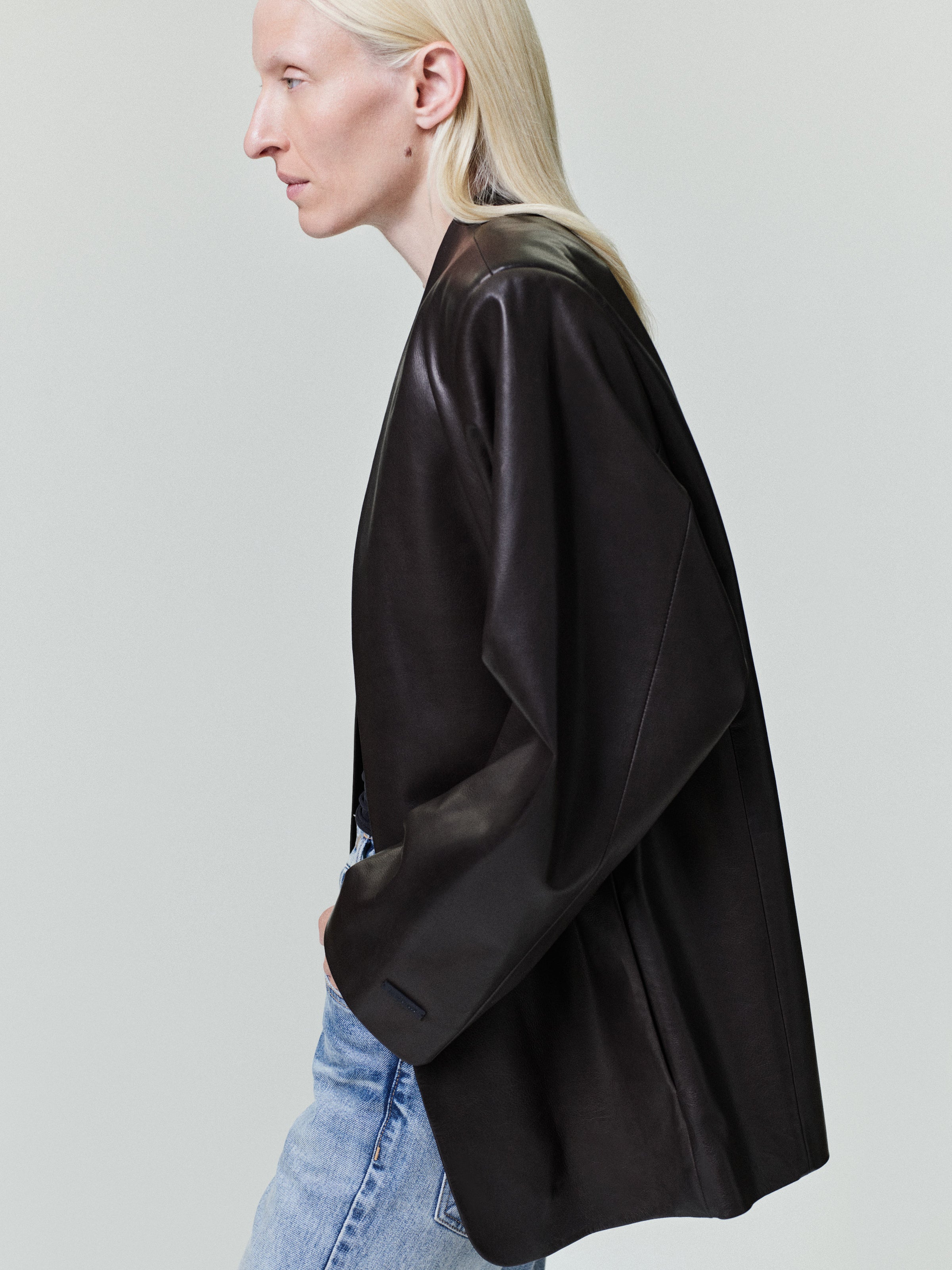 Side profile of a model wearing a dark Womens Leather Collarless Jacket paired with light blue jeans against a plain neutral background, highlighting the jacket's smooth texture and relaxed fit.