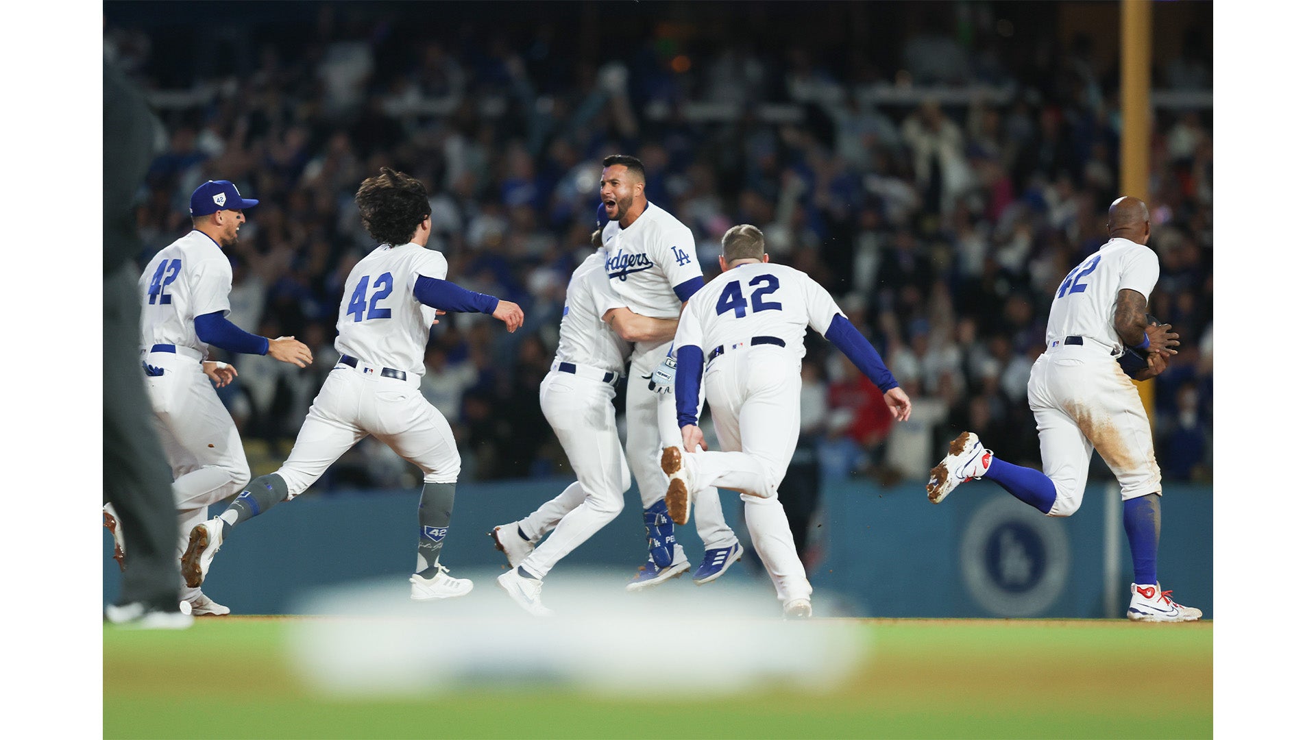 A baseball team dressed in white and blue uniforms celebrates on the field, running and jumping with excitement while a crowd watches from the stands behind them.