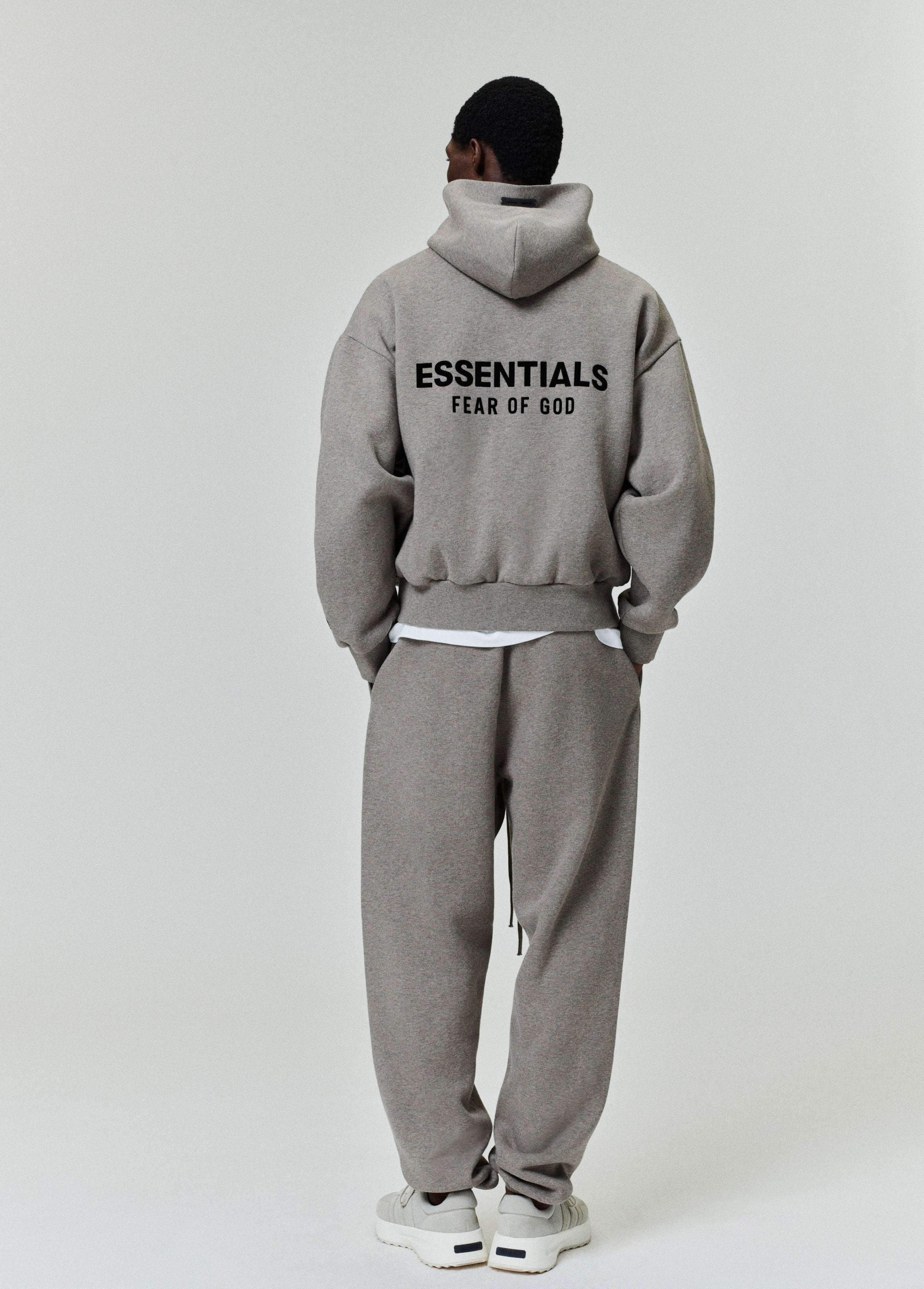 Rear view of taupe hoodie with bold black ESSENTIALS Fear of God logo across back paired with matching sweatpants