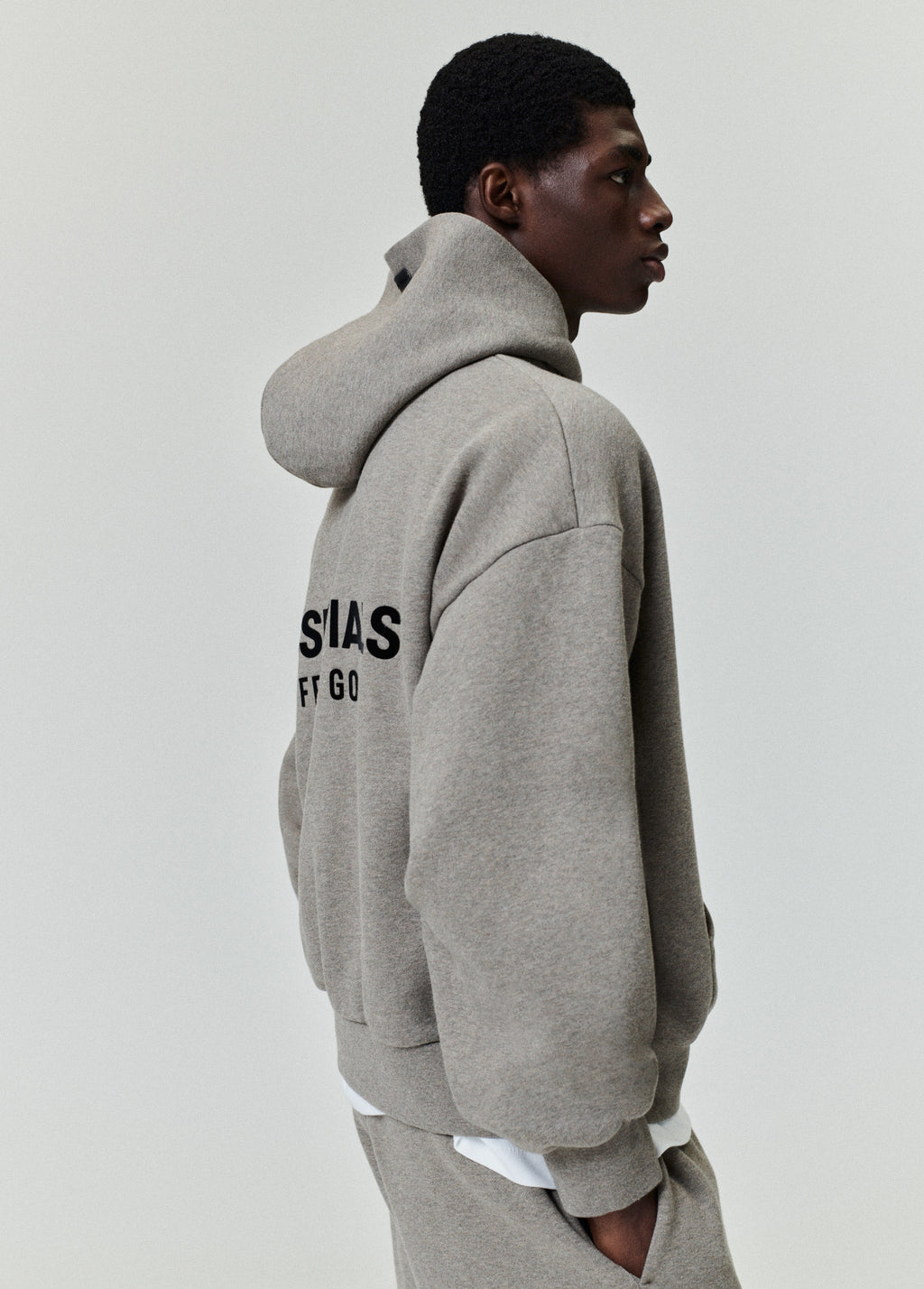Side profile of model wearing gray Classic Hoodie showing partial ESSENTIALS Fear of God logo on back, image A13_A11_3759.jpg