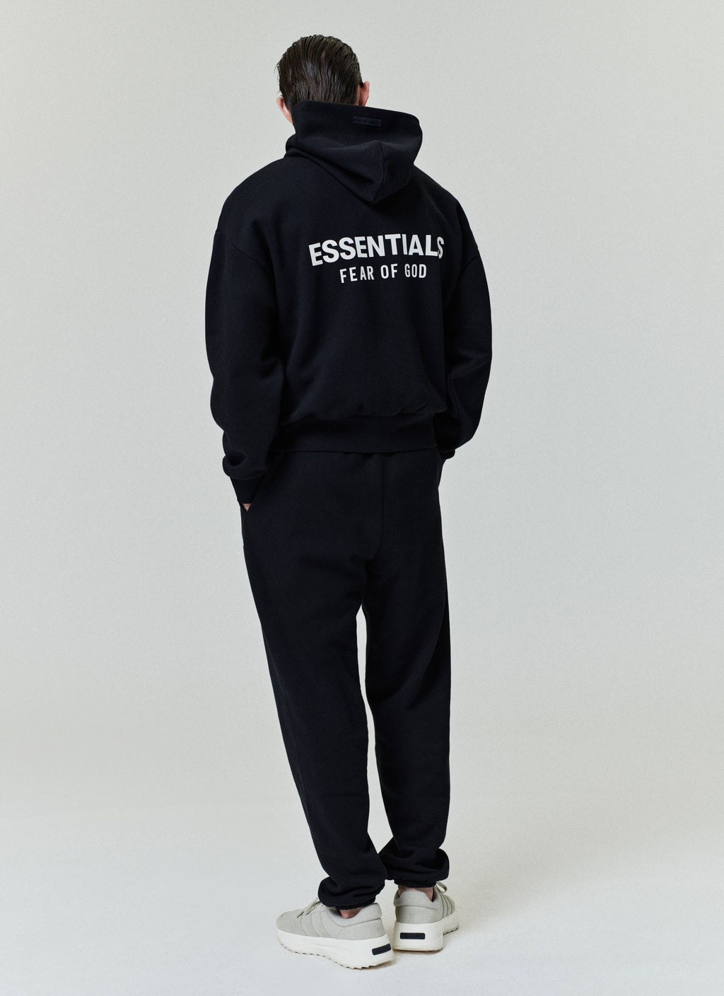 Black hoodie with hood and relaxed fit featuring large white "ESSENTIALS FEAR OF GOD" text on the back, paired with matching pants