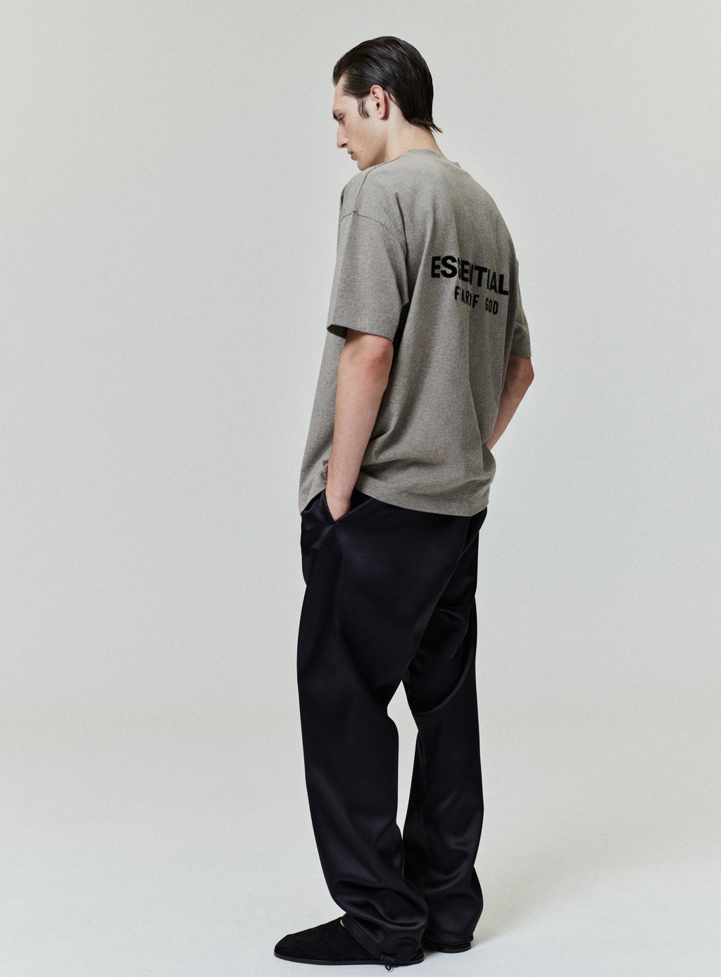 Side view of model wearing Homestead Heather Classic Short Sleeve Tee with bold ESSENTIALS Fear of God text on back, gallery image 0