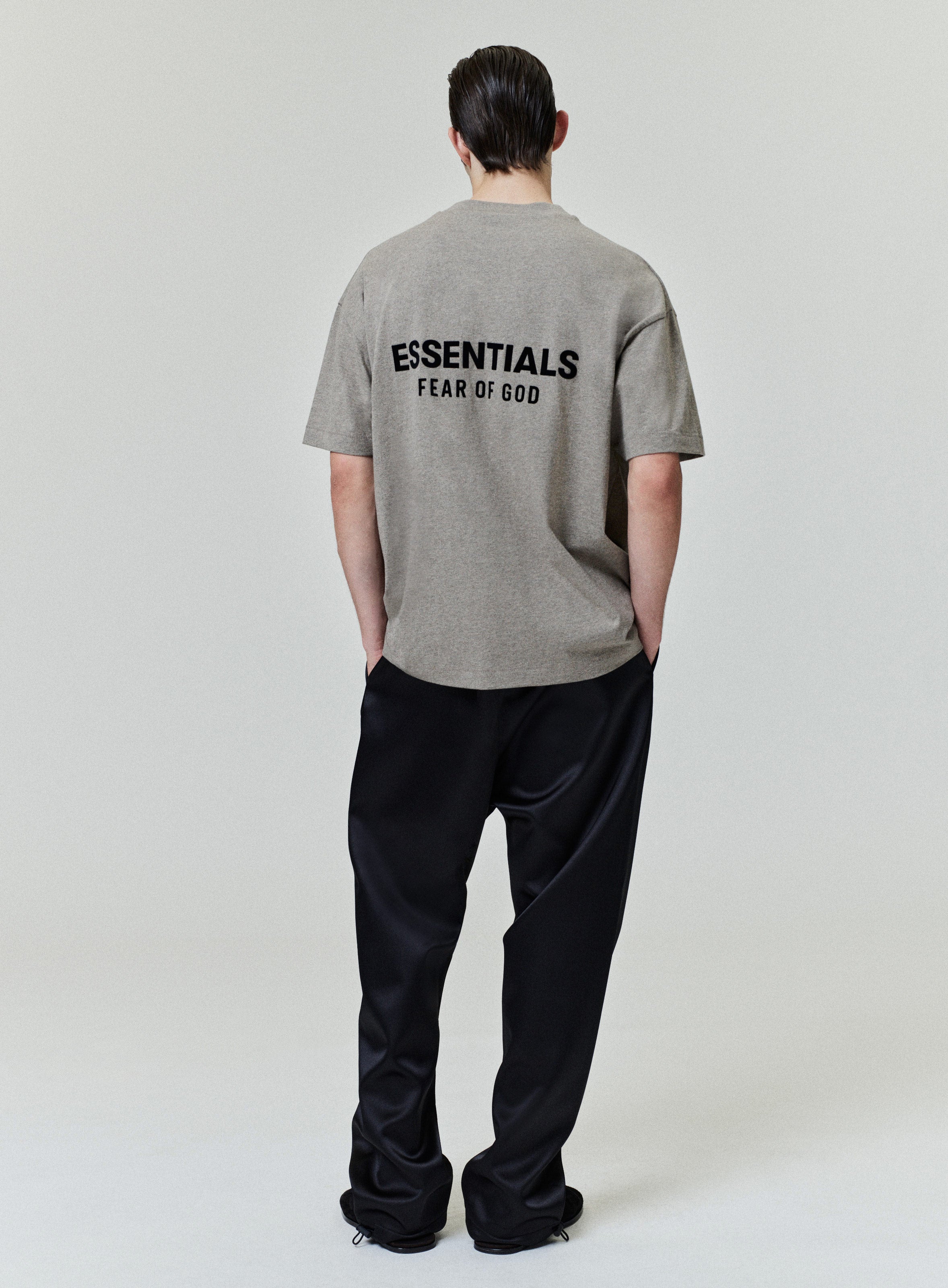 Model wearing taupe short sleeve tee with black ESSENTIALS Fear of God logo printed on upper back paired with black pants