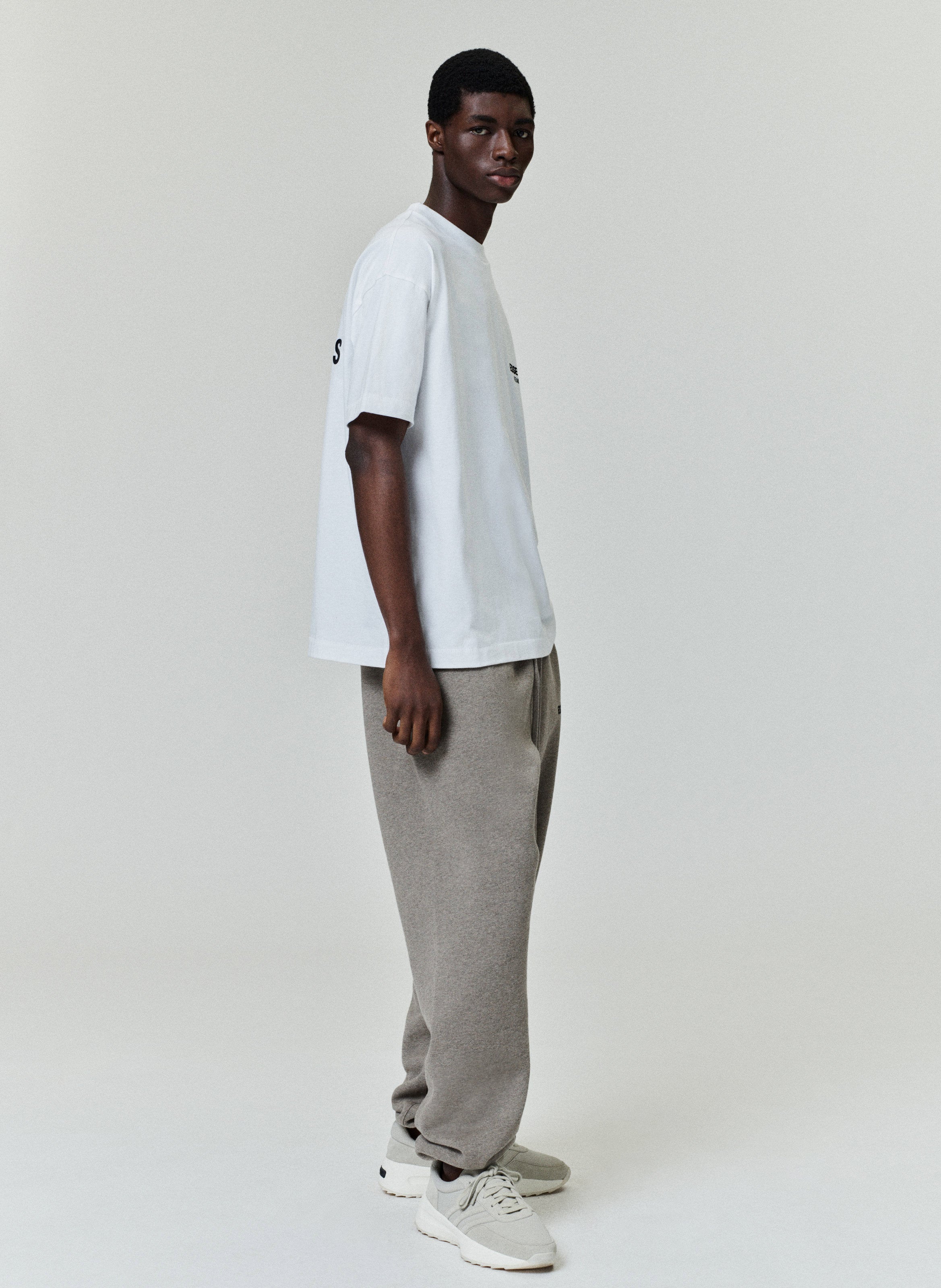 Model wearing a white Essentials Fear of God T-shirt with gray sweatpants against a light background.