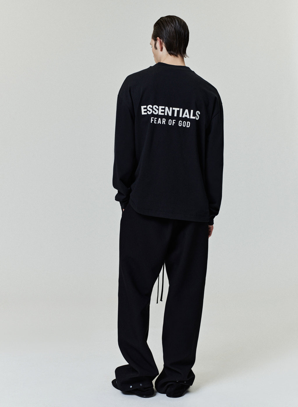 Rear view of model wearing black Classic Long Sleeve Tee with white ESSENTIALS Fear of God logo, gallery image 0