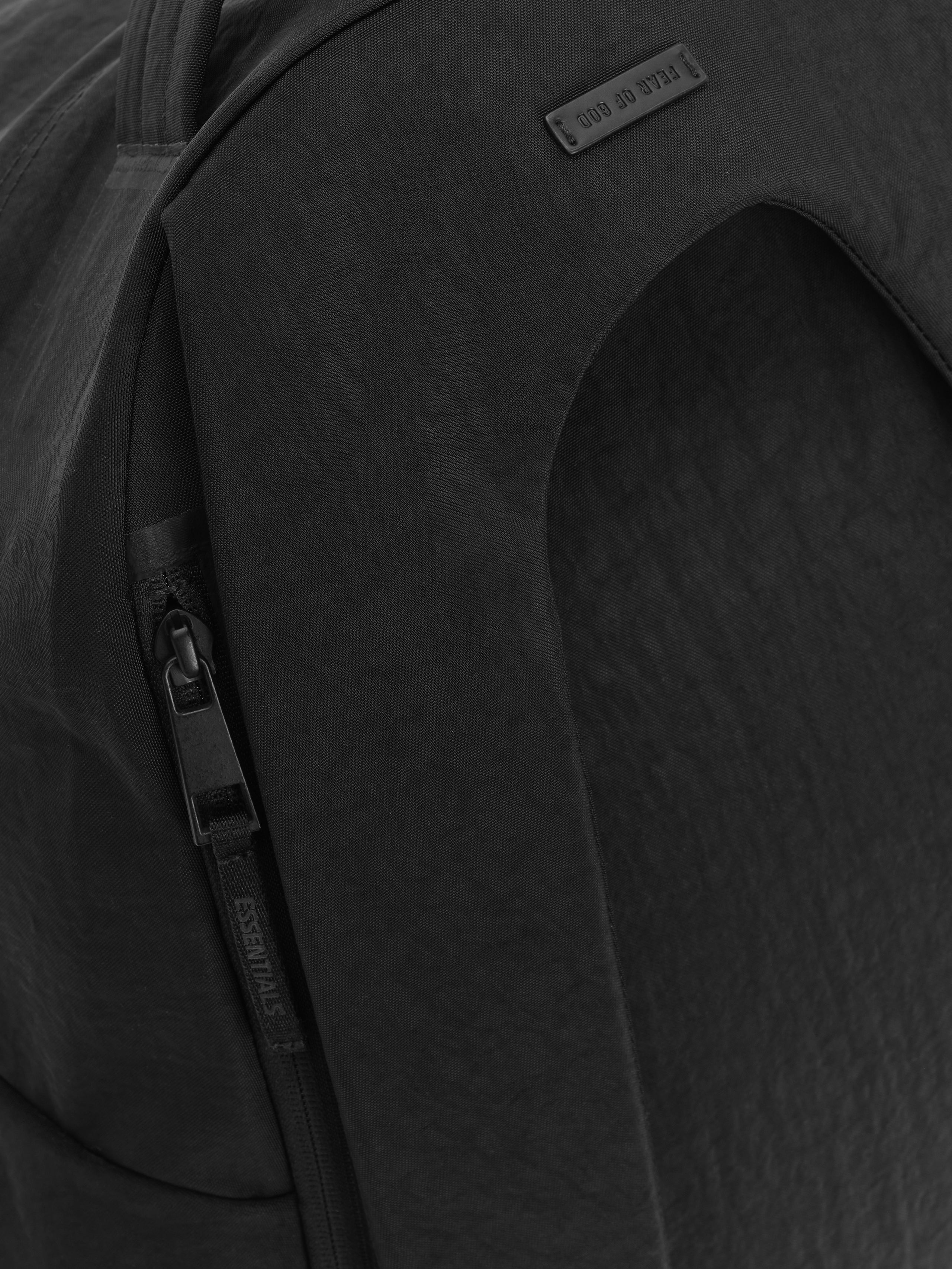 Essentials Backpack in black close up showing a curved front panel vertical zipper and embossed logo patch on textured fabric against a dark background