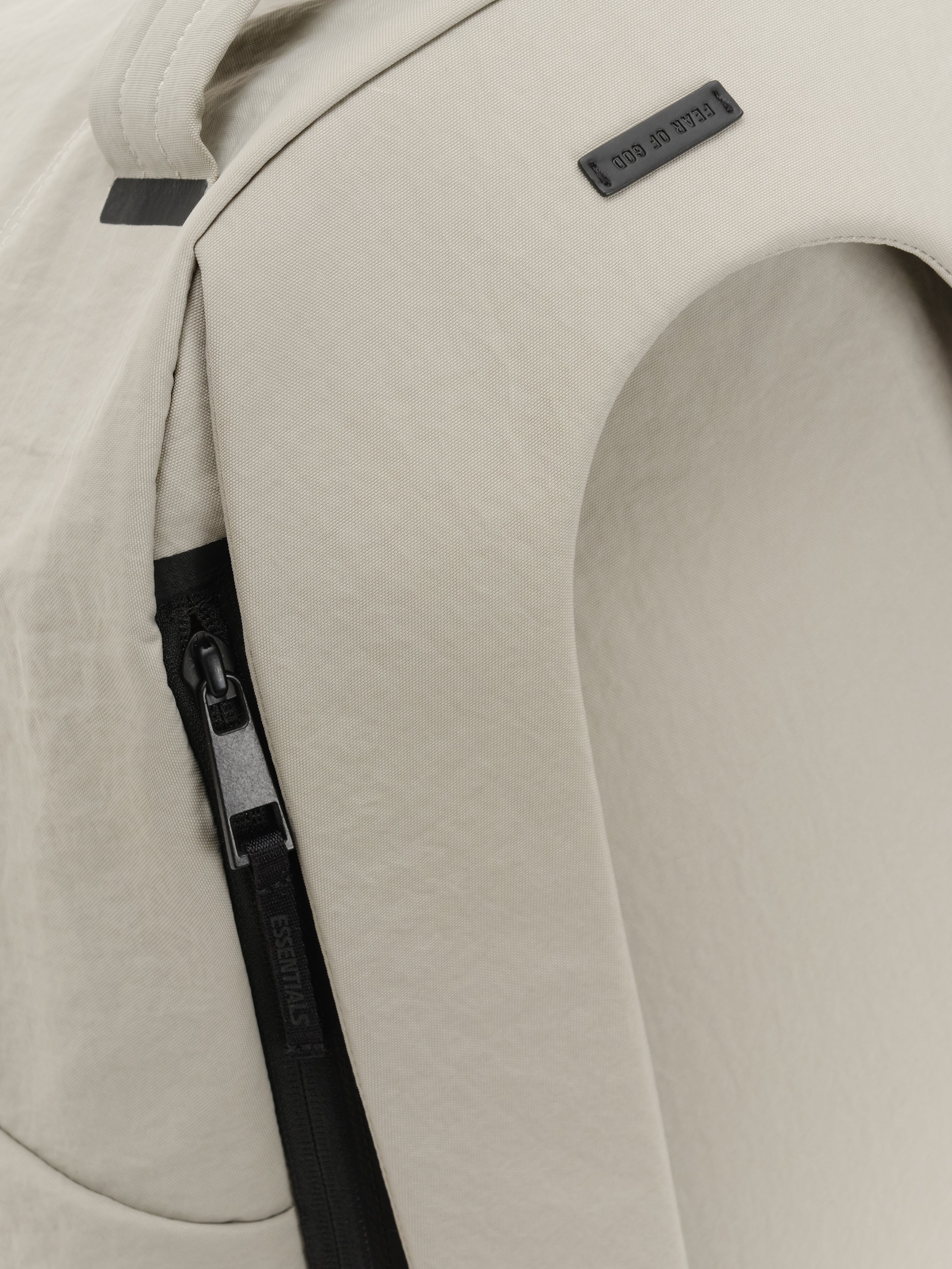 Essentials Backpack in beige close up showing a black side zipper padded shoulder strap and small rectangular logo patch on smooth textured fabric