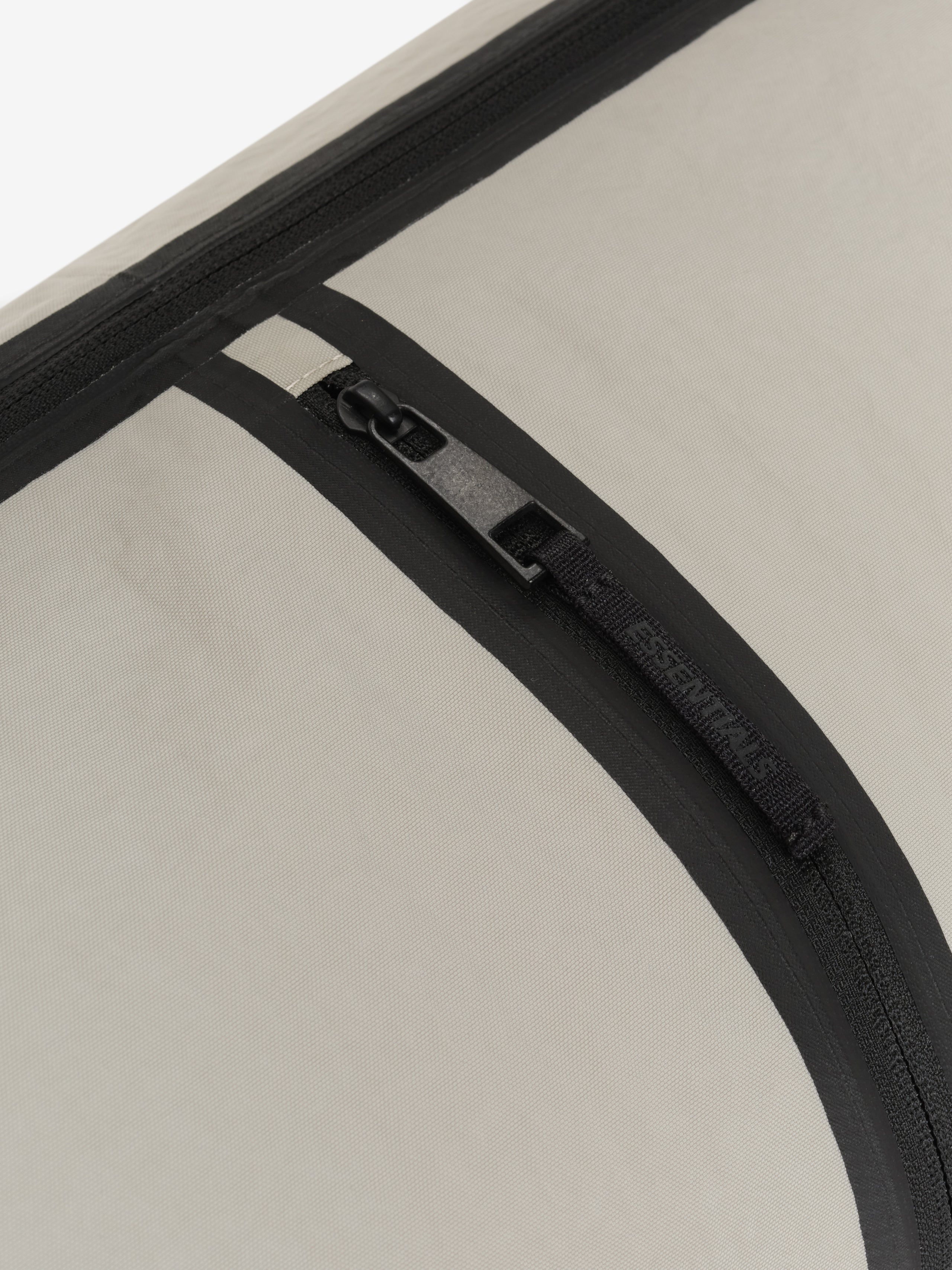 Essentials Shell Bag close up showing a black curved zipper with a metal pull and textured light beige fabric panel