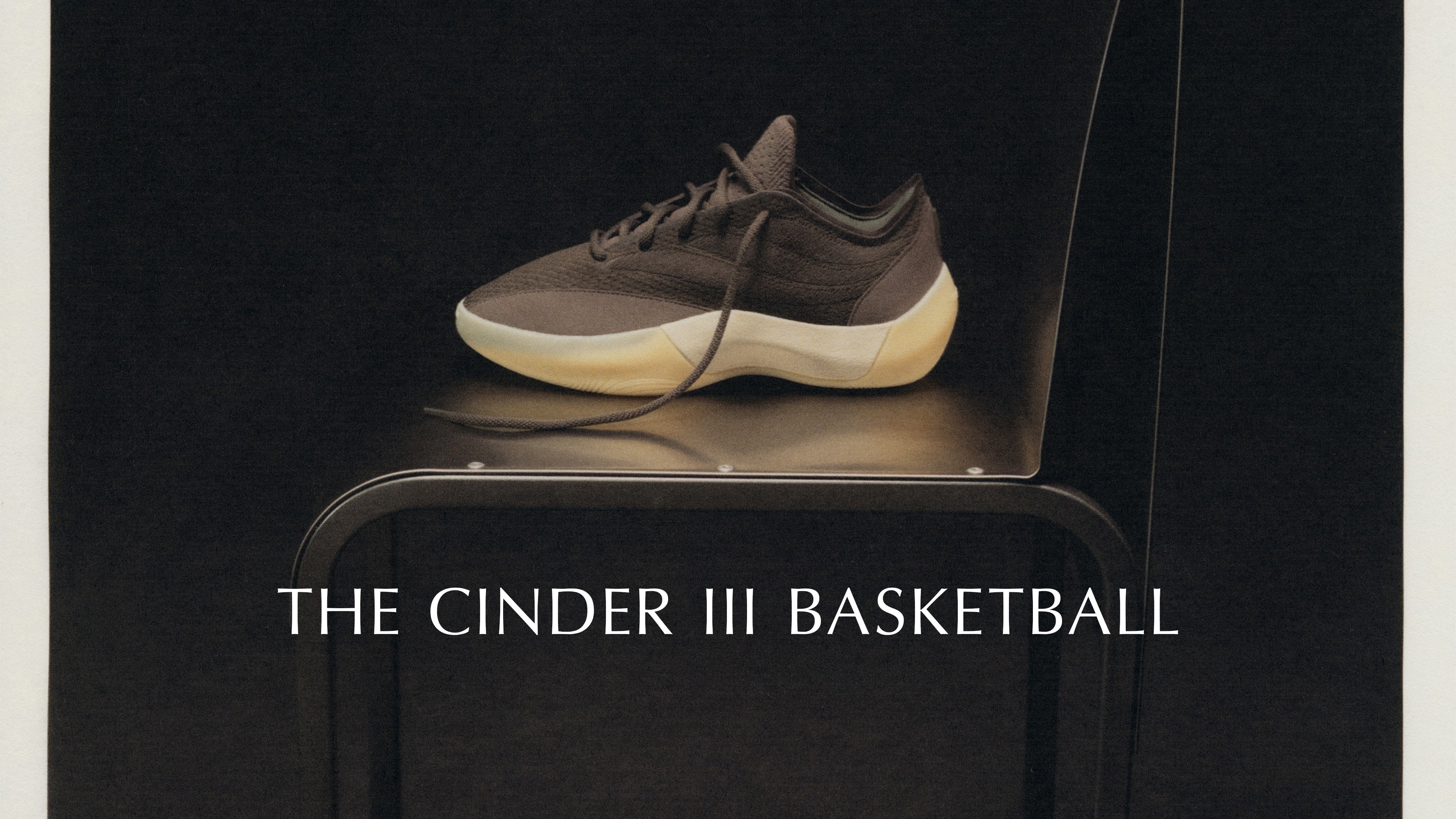 Side view of a sleek dark-toned basketball shoe with a thick white sole resting on a black chair against a dark background, accompanied by minimalist white text.